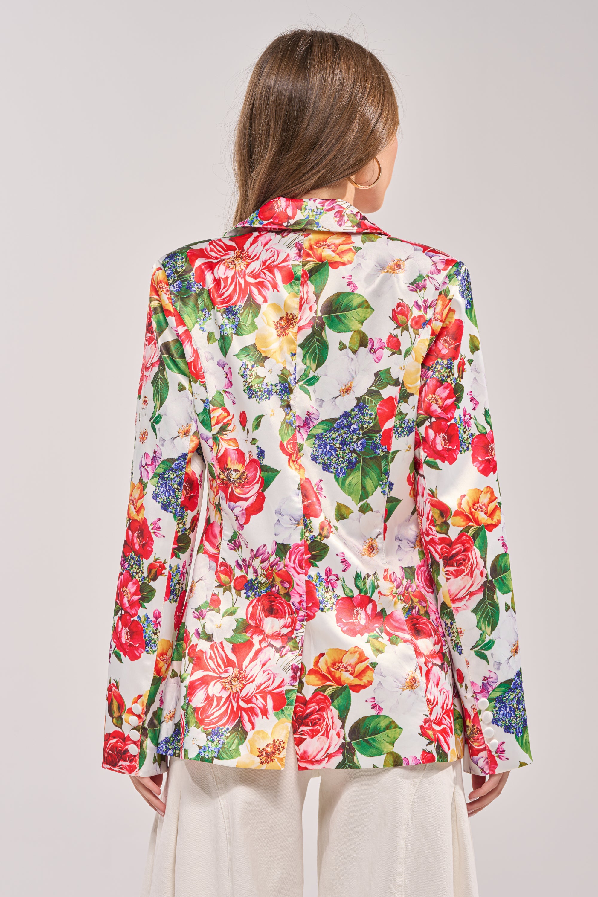 A woman faces away, wearing the ALESSANDRA SATIN BLAZER in vibrant red, pink, yellow, and green floral print over white pants. The tailored blazer stands out against a plain, light-colored background. 3