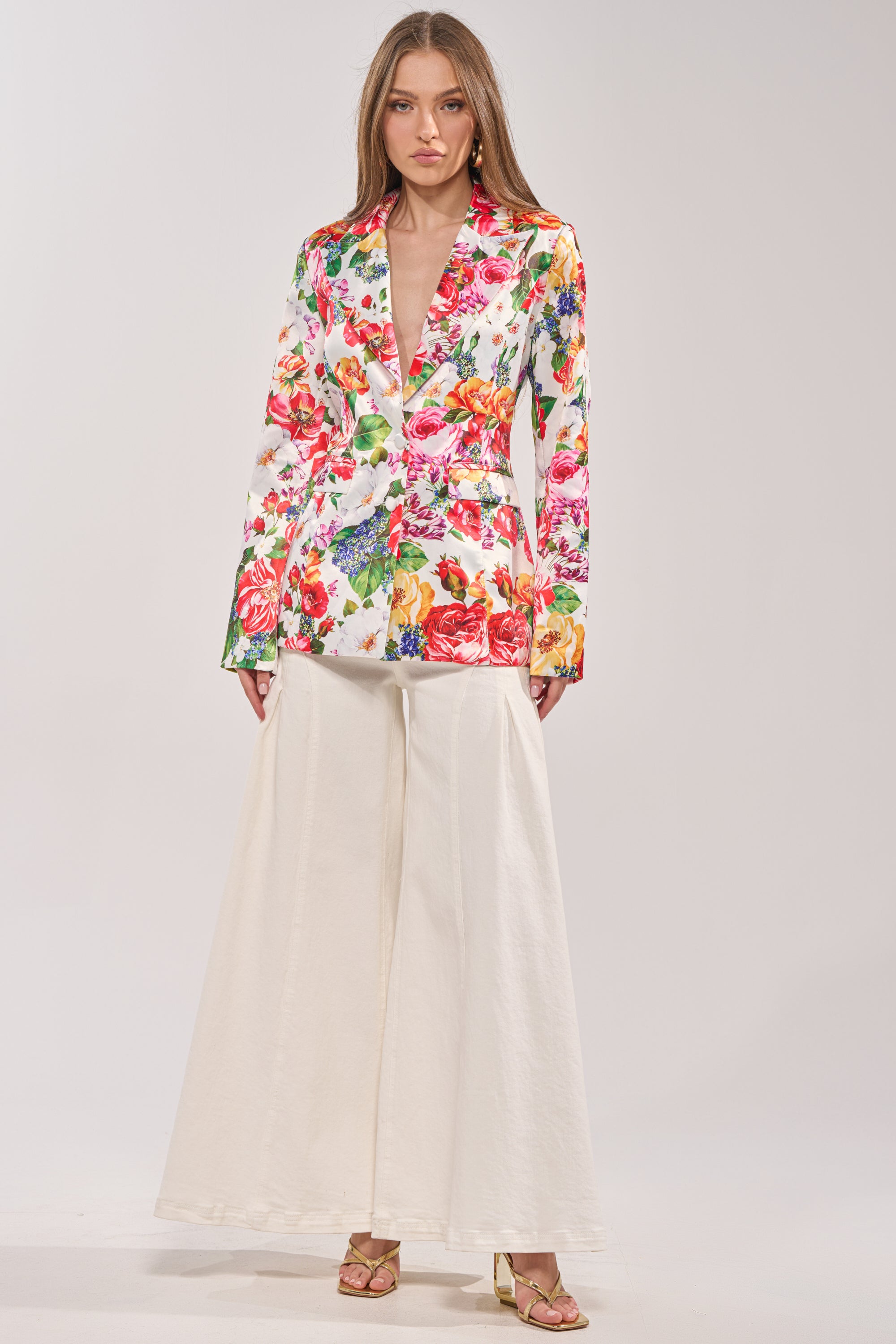 A woman models the ALESSANDRA SATIN BLAZER, featuring vibrant pink, red, and orange florals, styled with wide-leg cream trousers and gold strappy heels against a light background. 4
