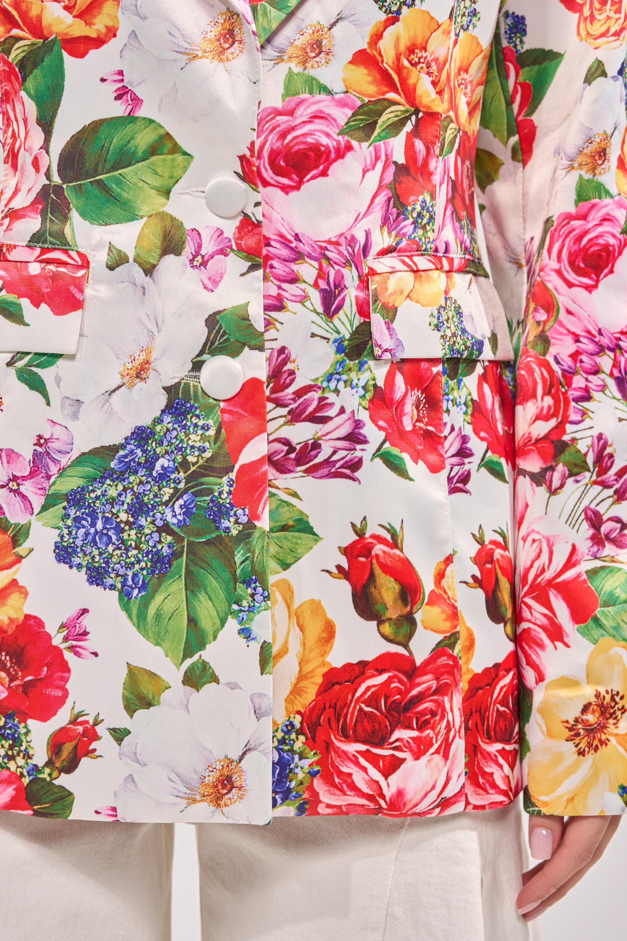 A close-up of a person wearing the ALESSANDRA SATIN BLAZER, featuring red, yellow, and white flowers with green leaves, white buttons, and flap pockets, paired with white pants. 5