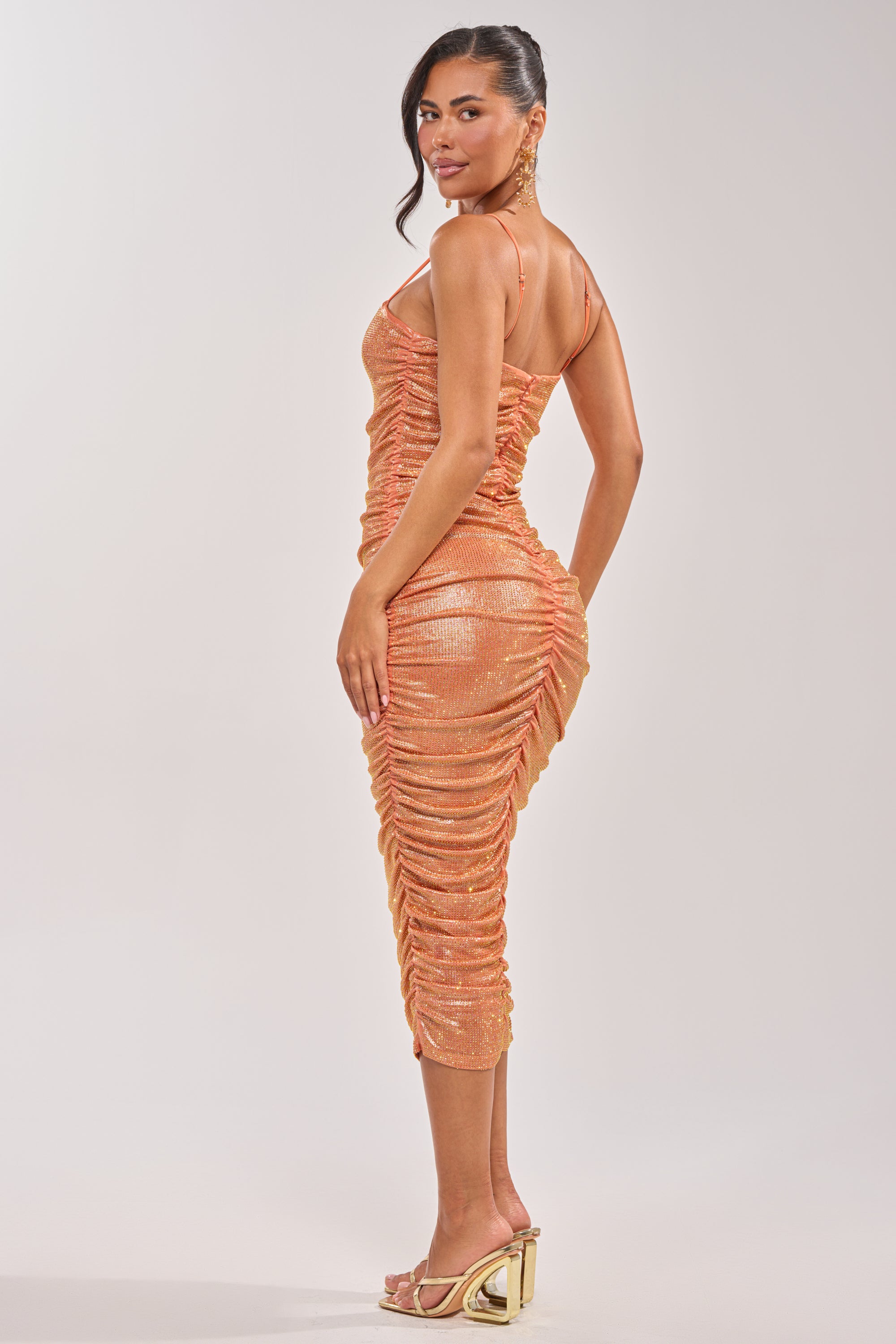 A woman poses in the KILL THE LIGHTS Strapless Rhinestone Midi Dress in Peach Bellini, standing sideways and looking over her shoulder against a plain background. 4