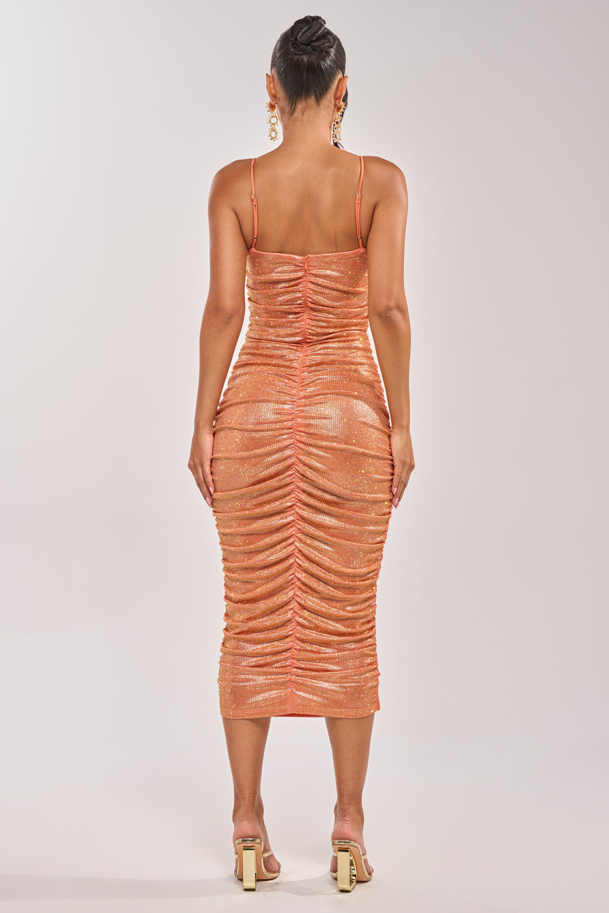 A woman faces away in the KILL THE LIGHTS STRAPLESS RHINESTONE MIDI DRESS IN PEACH BELLINI, paired with heels. Her hair is in a bun, and she wears dangling earrings against a plain light background. 5