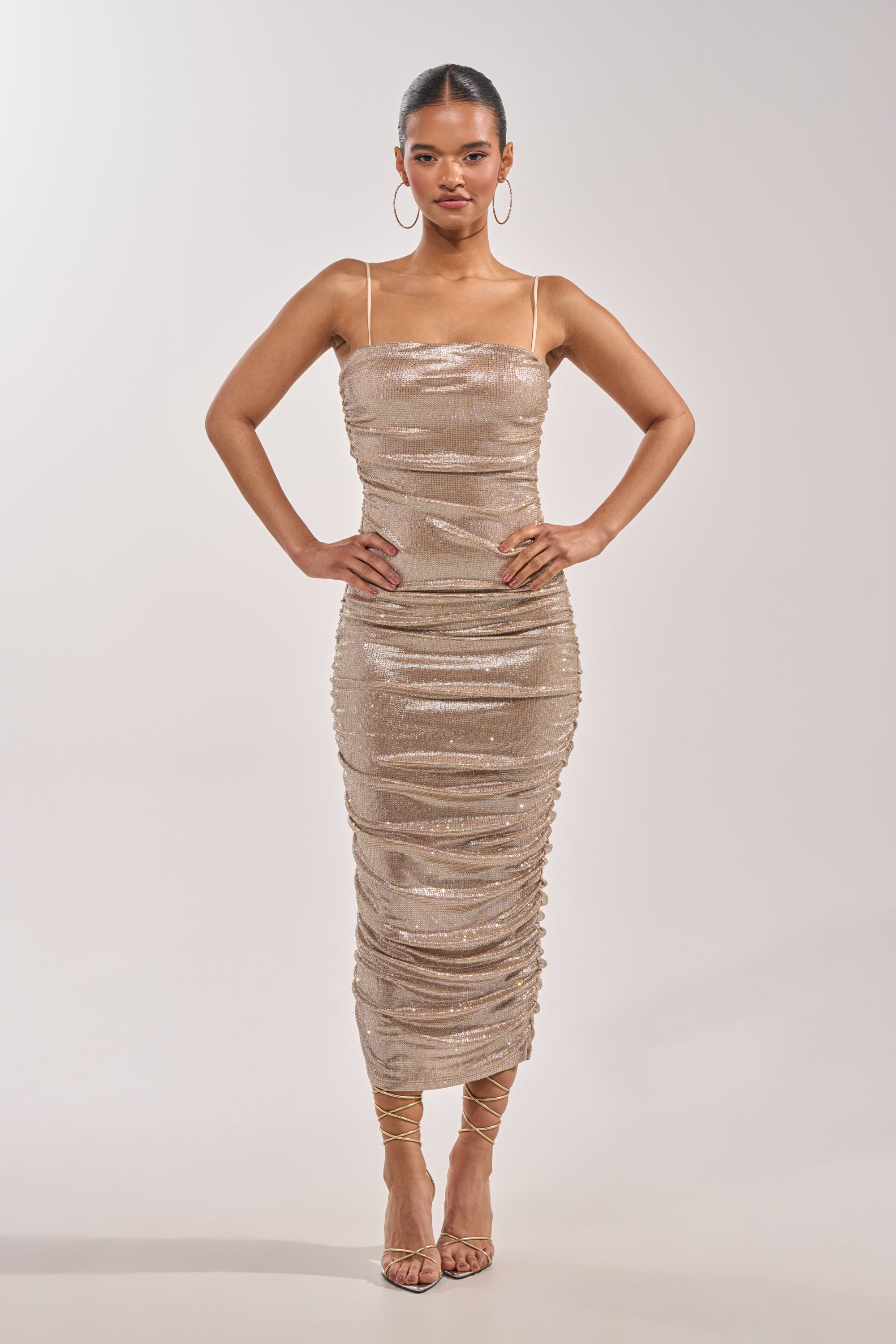 A woman poses confidently in the KILL THE LIGHTS STRAPLESS RHINESTONE MIDI DRESS IN CHAMPAGNE, featuring gold sequins and ruching with a mid-calf fit. She accessorizes with hoop earrings and strappy heels, hair slicked back, against a plain light background. 2