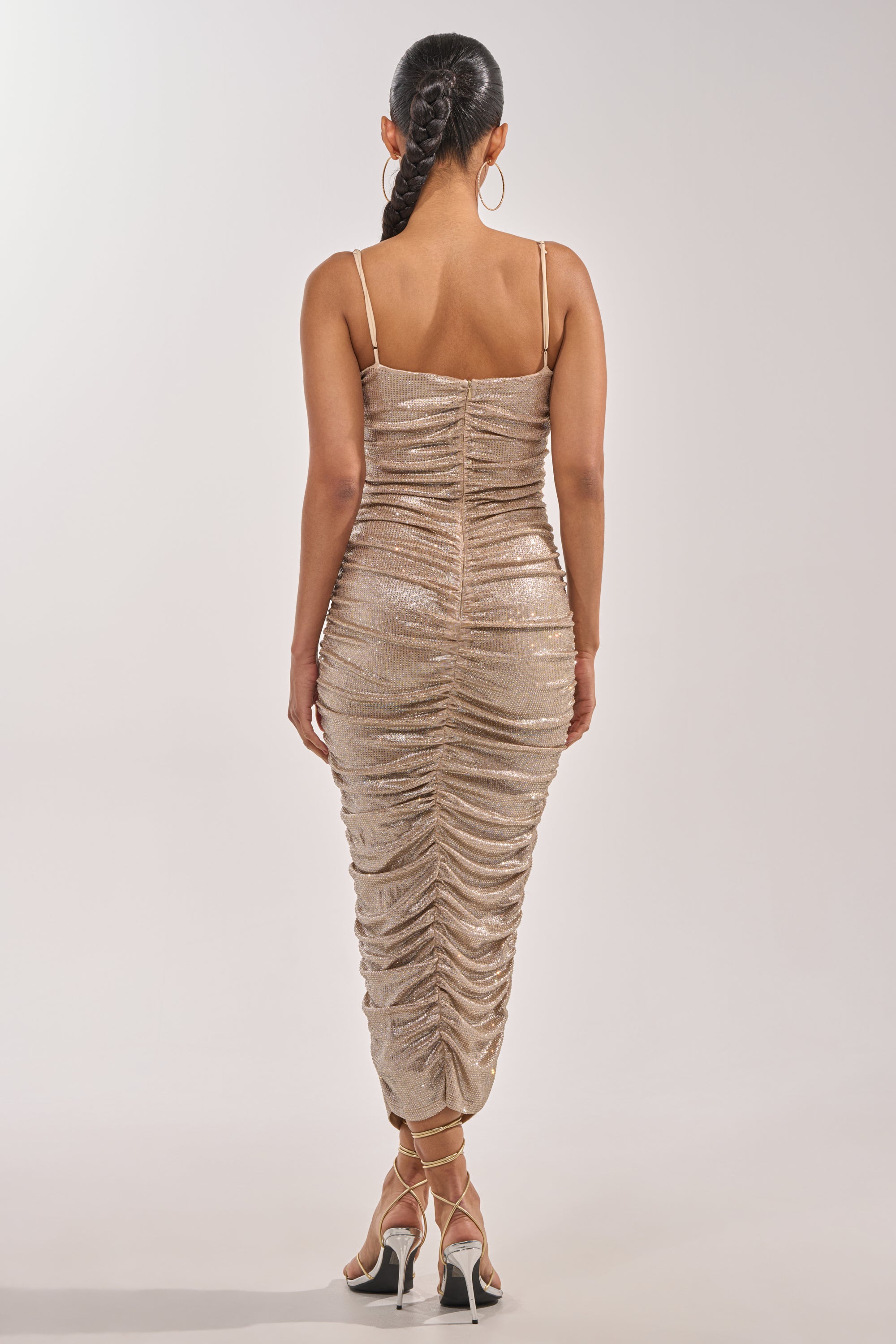 A woman with braided hair wears the KILL THE LIGHTS STRAPLESS RHINESTONE MIDI DRESS IN CHAMPAGNE and high heels, standing against a plain white background, facing away from the camera. 4