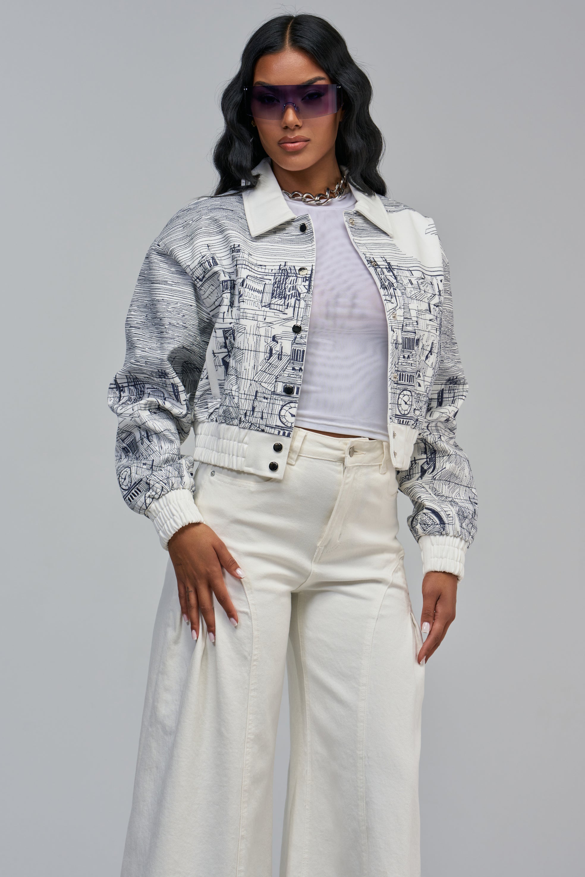 A person wears a white CATCH MY DRIFT EMBROIDERED BOMBER jacket with navy blue line-drawing illustrations, a white top, and wide-leg white pants.