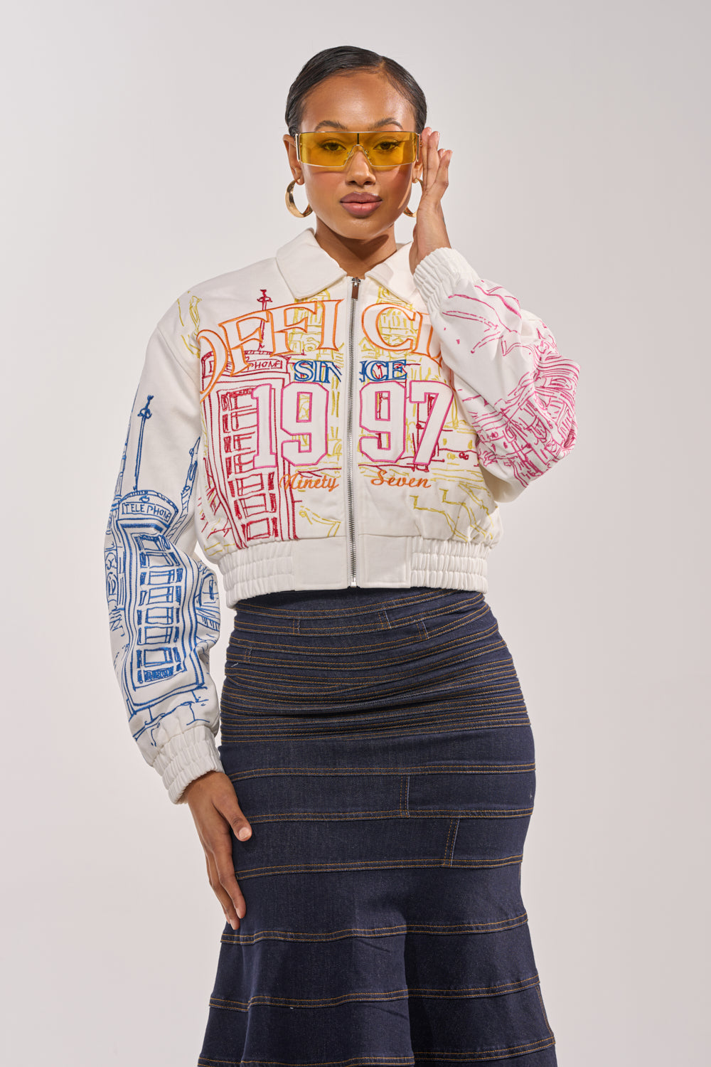 A woman stands confidently against a plain light background, wearing yellow-tinted sunglasses and the BEEN AROUND THE BLOCK EMBROIDERED BOMBER, styled with a dark, striped flared skirt. 0