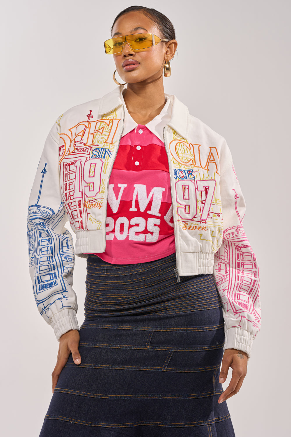 A woman exudes sporty-chic style in yellow sunglasses, a pink “WVM 2025” polo, dark denim skirt, and the BEEN AROUND THE BLOCK EMBROIDERED BOMBER with multicolored details against a plain background. 1