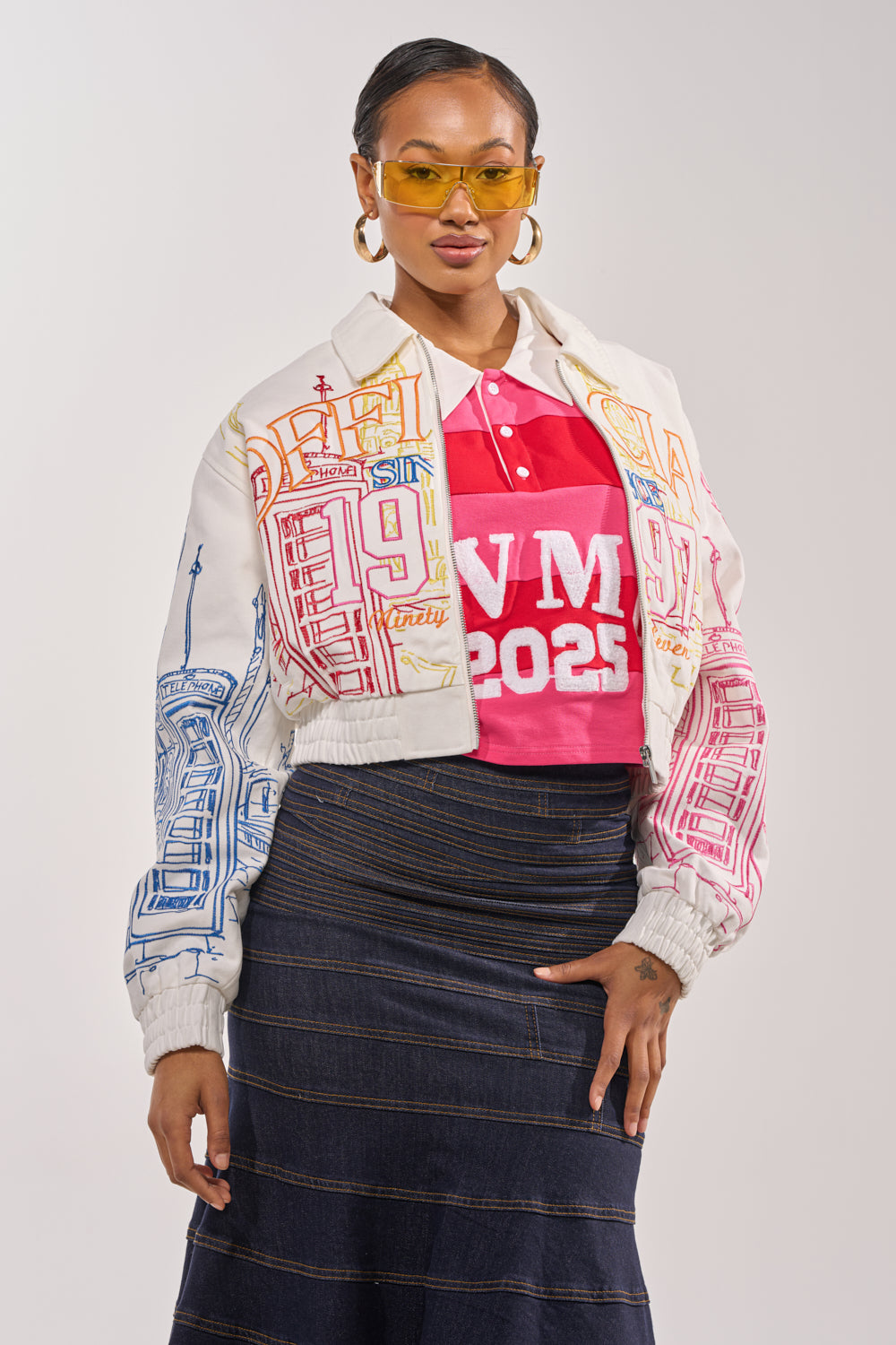 A woman sports yellow sunglasses, a red “NM 2025” shirt, the BEEN AROUND THE BLOCK EMBROIDERED BOMBER with cityscape graphics, and a dark denim skirt, exuding bold street style against a plain background. 2