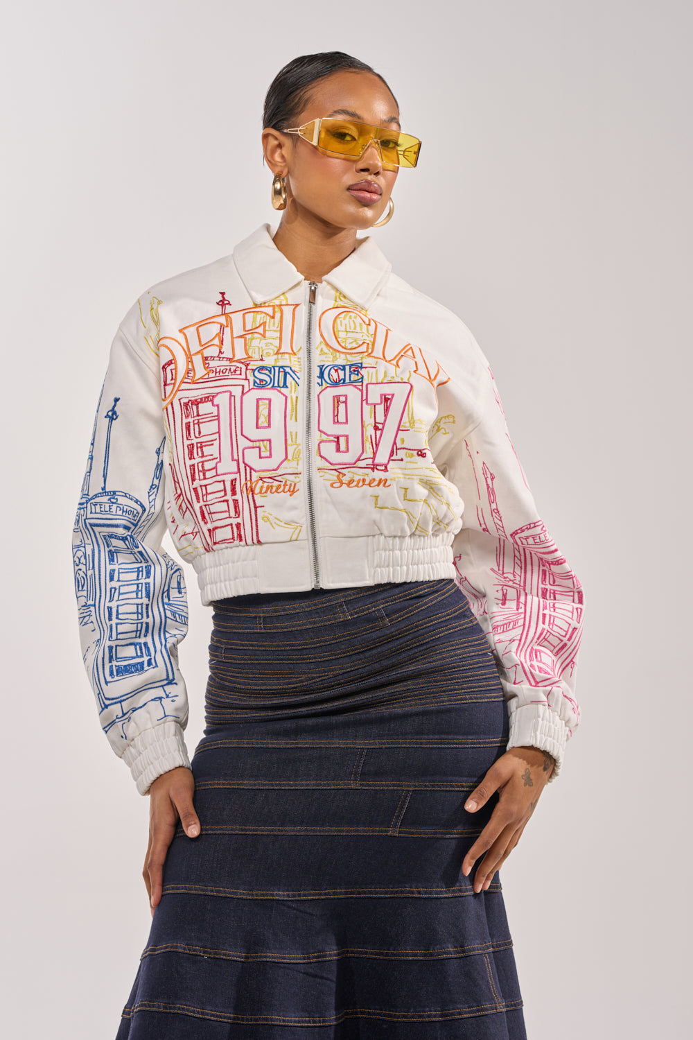 A woman poses confidently in yellow sunglasses, a dark denim skirt, and the BEEN AROUND THE BLOCK EMBROIDERED BOMBER with cityscape graphics and "1997" text for a chic street-style look. 3