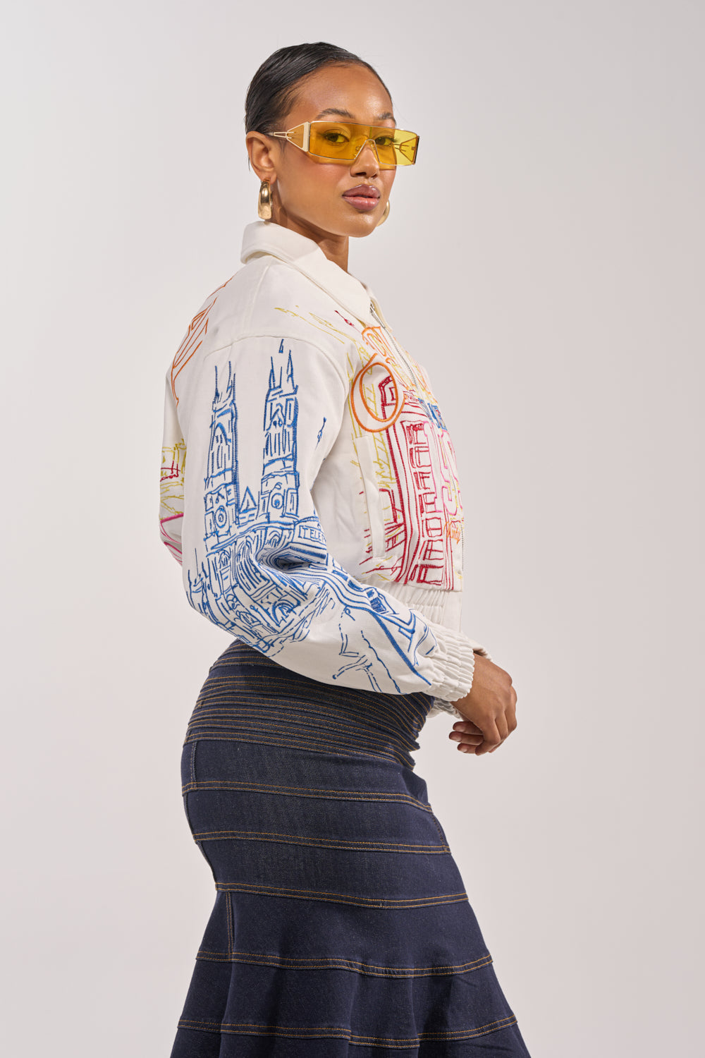 A woman poses sideways against a plain light background, wearing yellow-tinted sunglasses and the BEEN AROUND THE BLOCK EMBROIDERED BOMBER over a dark striped skirt. 4