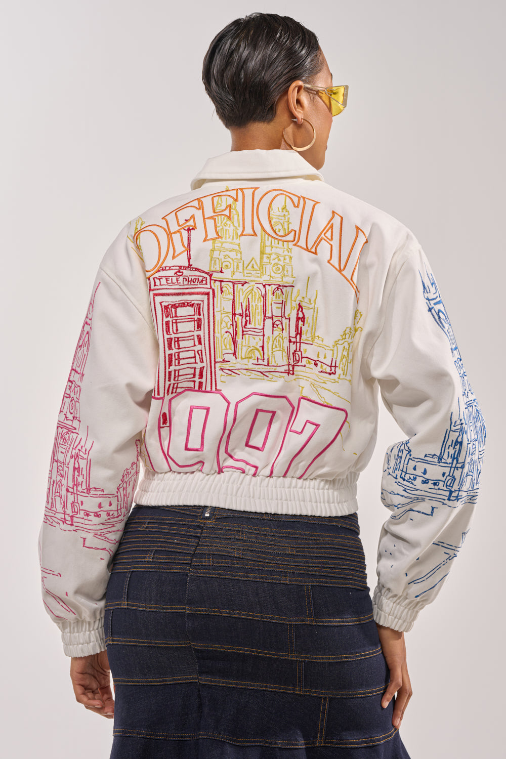 A person with short hair and yellow sunglasses is seen from behind wearing the BEEN AROUND THE BLOCK EMBROIDERED BOMBER, a white jacket with colorful cityscape graphics and “OFFICIAL 1997” on the back, paired with a dark denim skirt. 6
