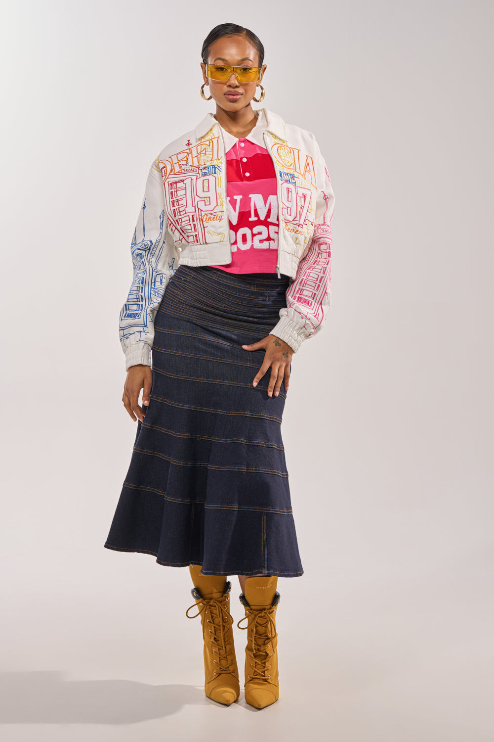 A woman poses confidently in yellow sunglasses, a red "W M 2025" shirt, a navy flared skirt, tan lace-up boots, and the BEEN AROUND THE BLOCK EMBROIDERED BOMBER against a plain background. 7