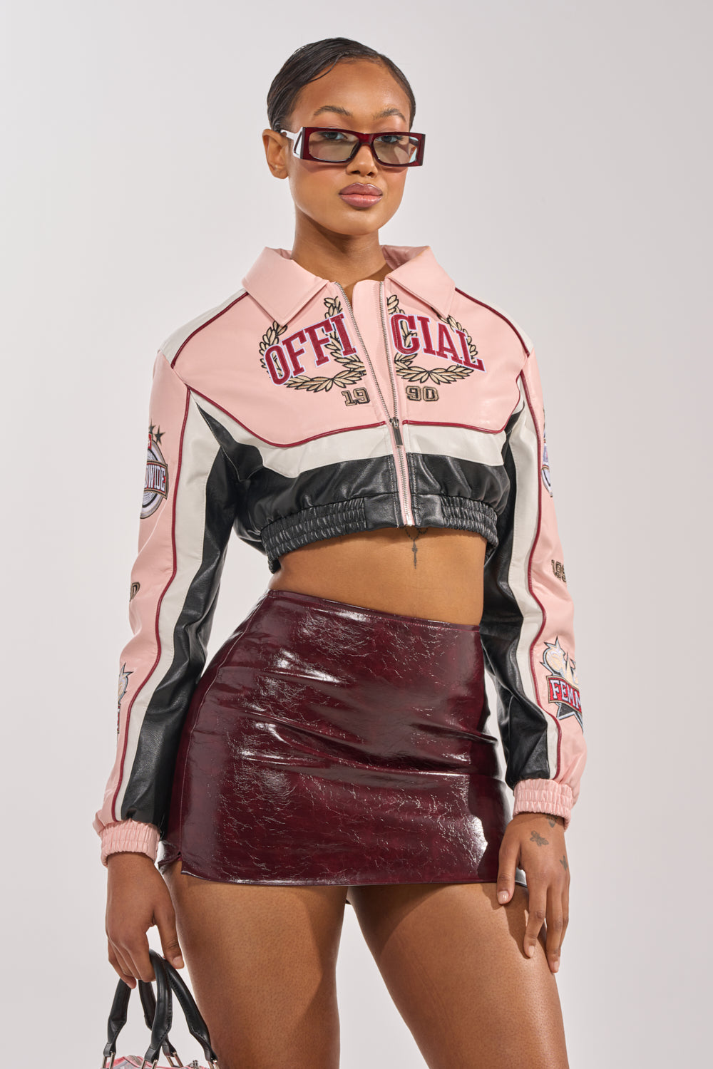 A woman wearing sunglasses, the NEVER BETTER CROPPED BOMBER with varsity-style patches, and a shiny dark red mini skirt stands holding a small bag against a plain background. 0