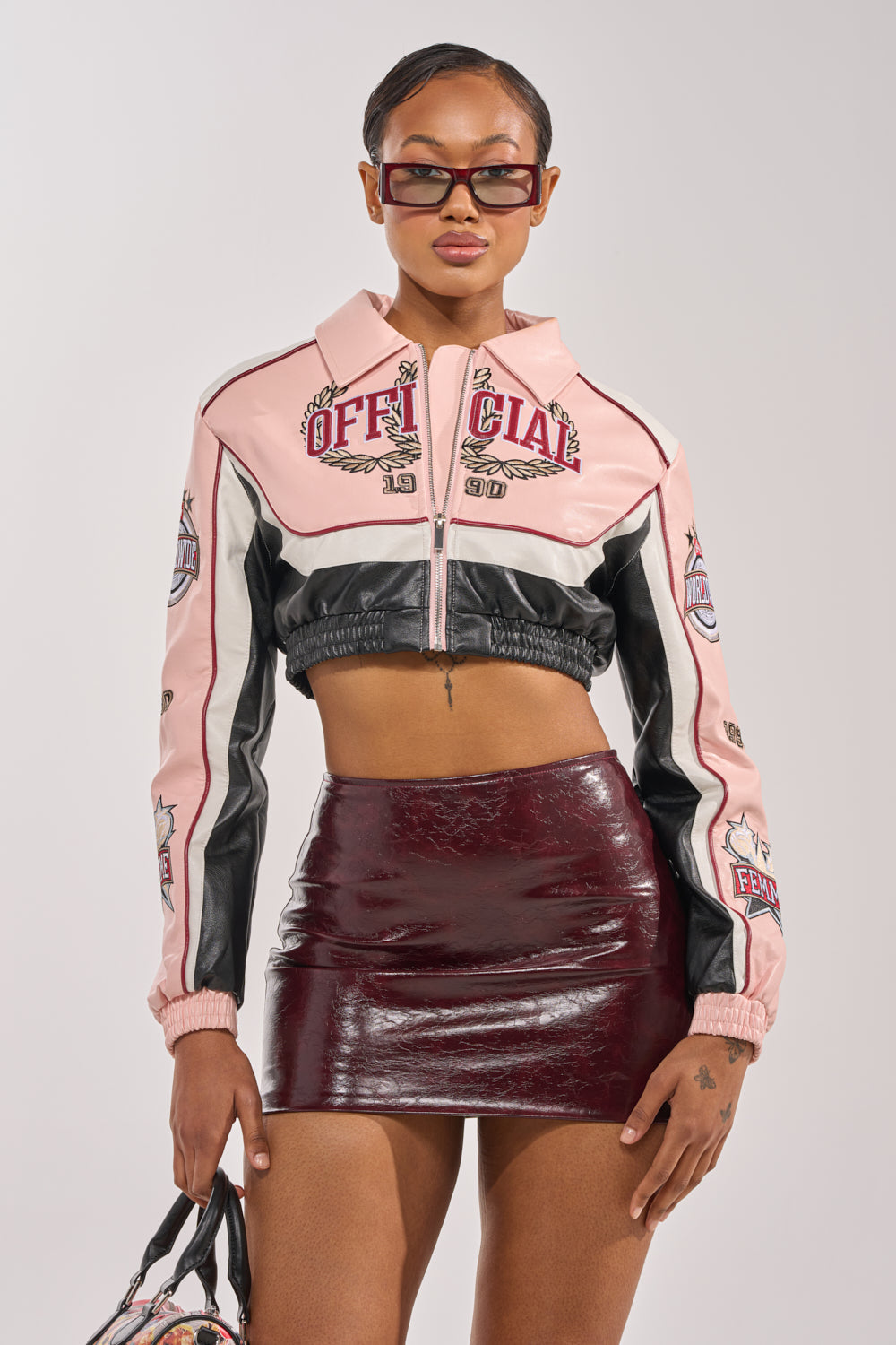 A woman in sunglasses and the NEVER BETTER CROPPED BOMBER jacket with "Official" patches, a shiny burgundy mini skirt, and matching handbag stands facing forward. The background is plain and light-colored. 1