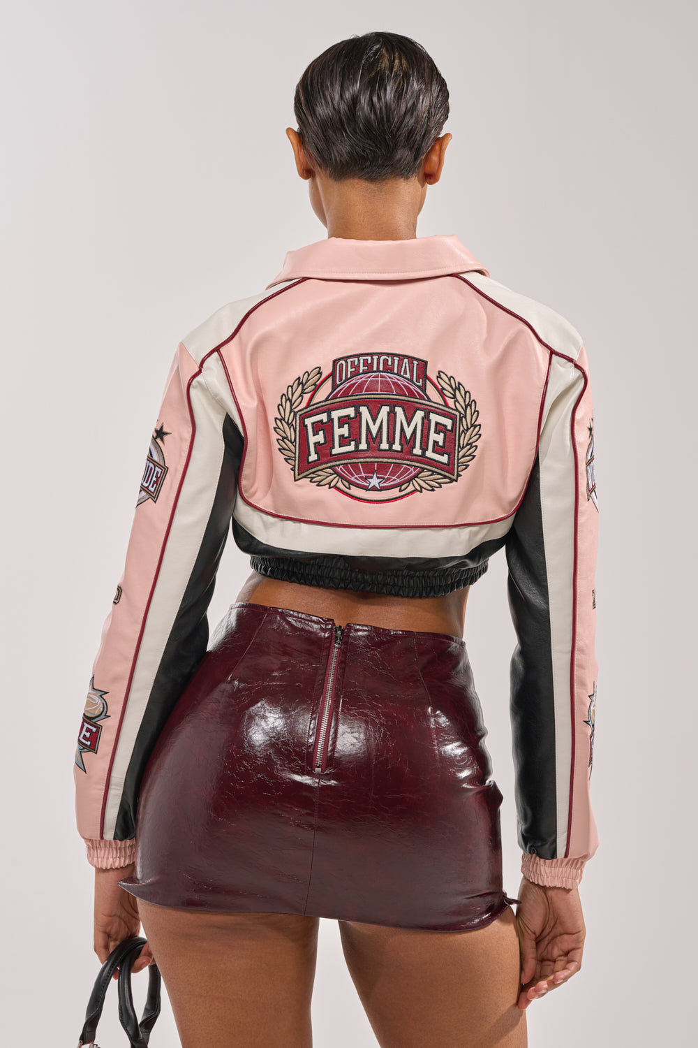 A person stands facing away, wearing the NEVER BETTER CROPPED BOMBER in pink faux leather with "Official Femme" on the back and a shiny burgundy mini skirt, holding sunglasses in their left hand. 3