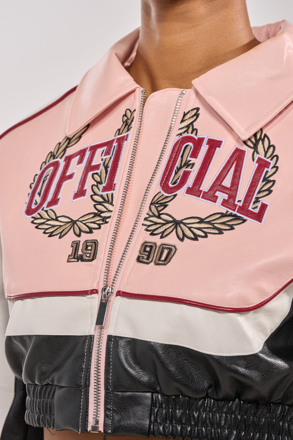 A close-up of a person wearing the NEVER BETTER CROPPED BOMBER, a black and pink faux leather jacket with "OFFICIAL" and "1990" embroidered on the chest, laurel wreath accents, a zipper, and a light pink collar. 6