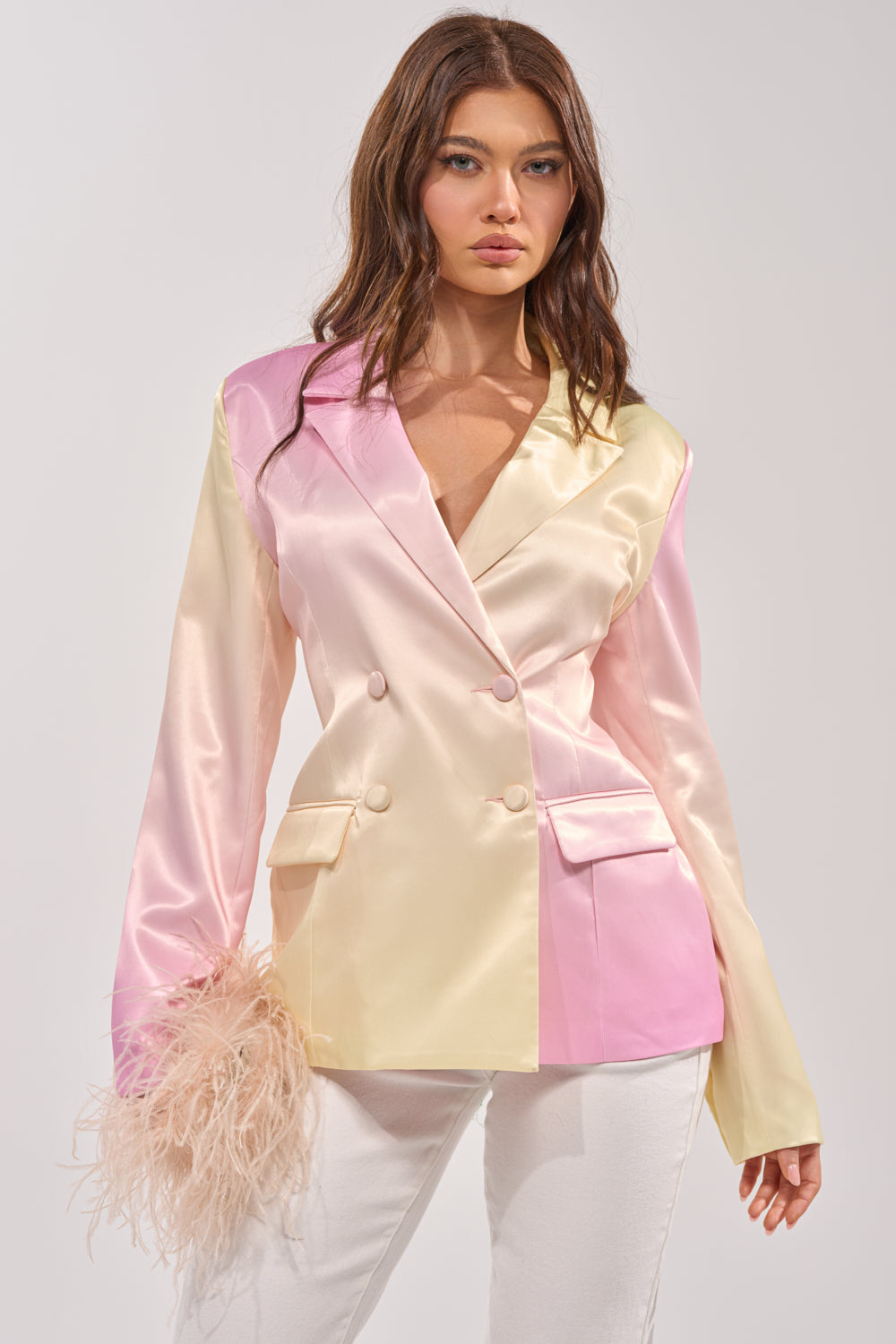 A woman wears the SWEET TALK SATIN BLAZER, a glossy double-breasted piece in pink and cream hues, paired with white pants. She holds a fluffy blush accessory and stands against a simple light backdrop. 0