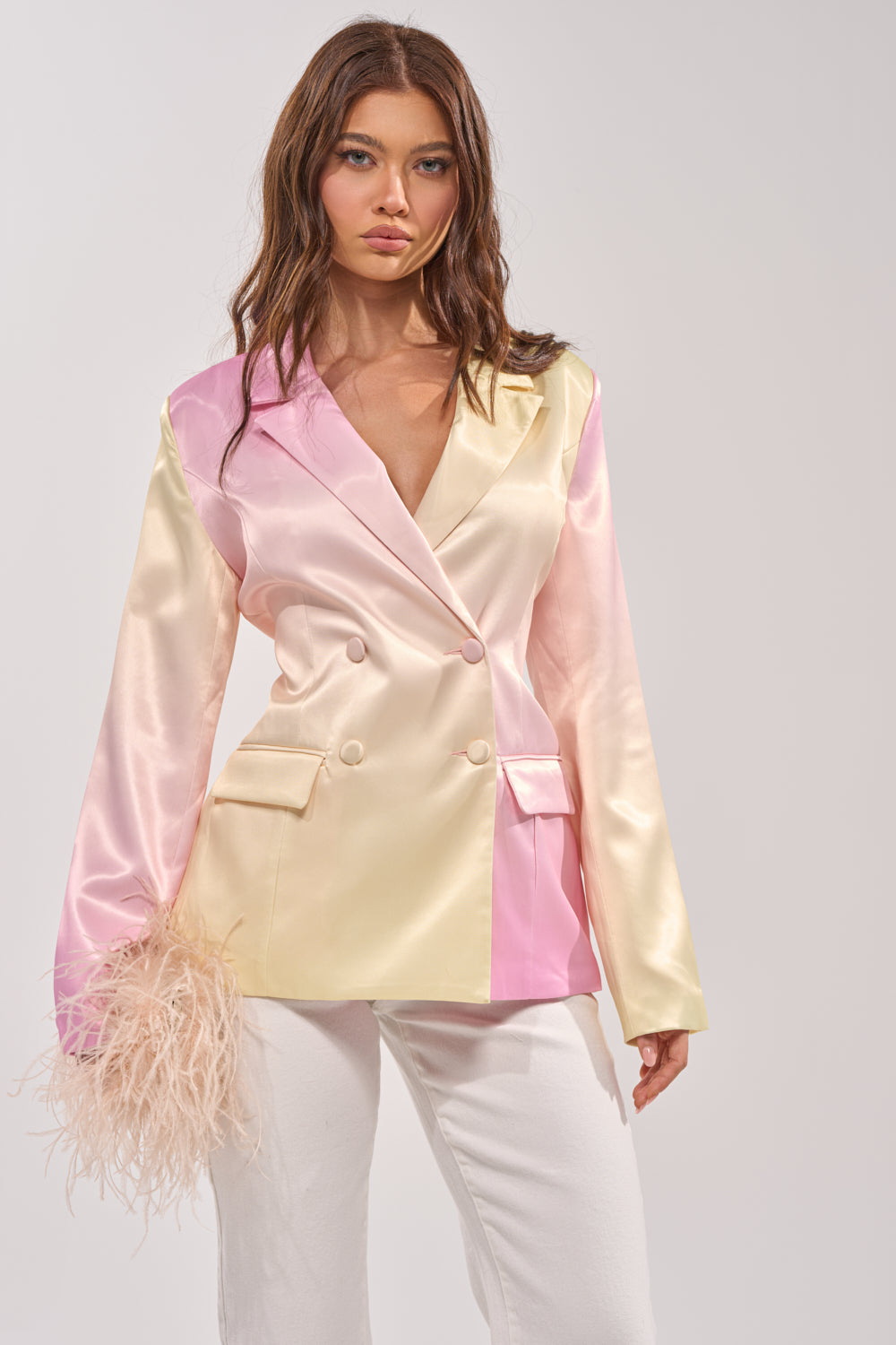 A woman models the SWEET TALK SATIN BLAZER in pastel pink and yellow blocks, paired with white pants and holding a beige feather accessory, standing against a plain light background. 1