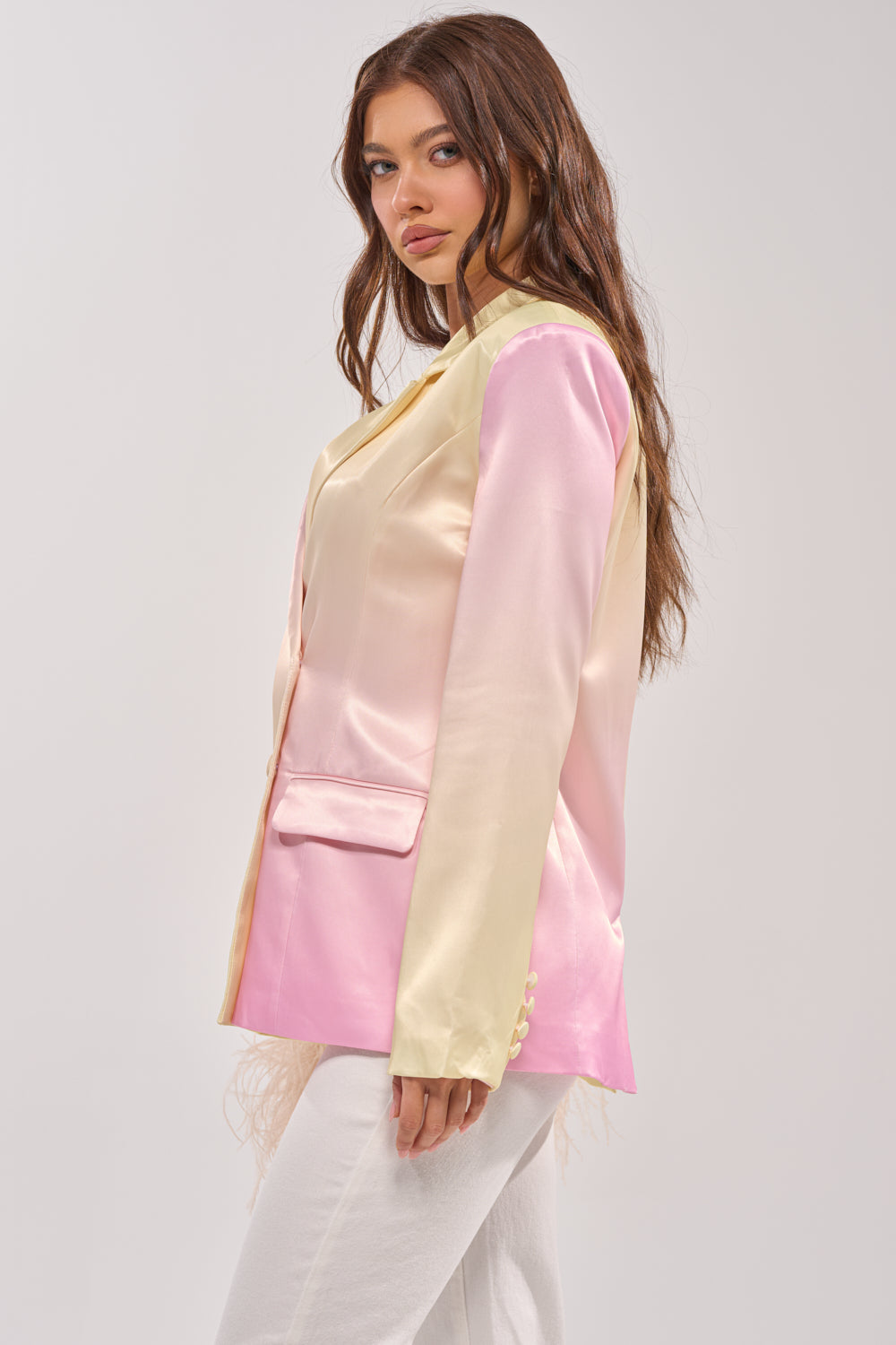 A woman with long brown hair models the SWEET TALK SATIN BLAZER in shiny pastel pink and yellow, paired with white pants and shown in side profile against a plain light gray background. 2