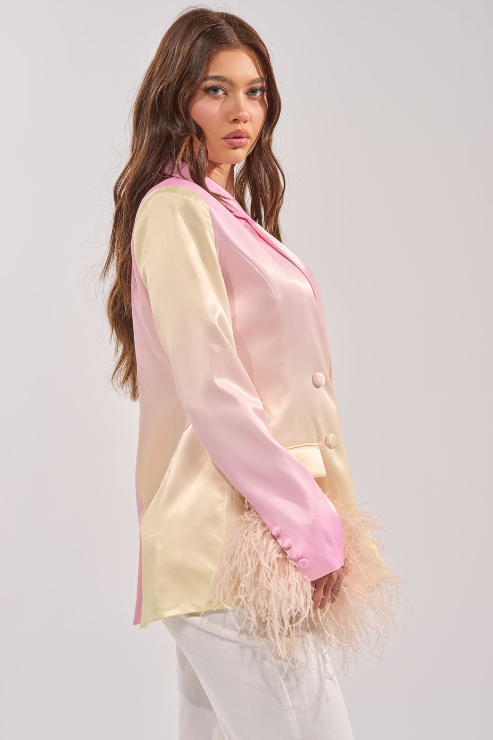 A woman with long wavy brown hair wears the SWEET TALK SATIN BLAZER with feather-detailed sleeves, paired with white pants, standing against a plain light background. 3