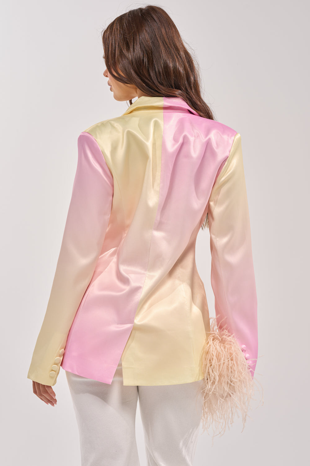 A woman with long brown hair models the SWEET TALK SATIN BLAZER in yellow and blush pink tones, seen from the back. The tailored blazer features a feather detail on one sleeve and is paired with crisp white pants. 4