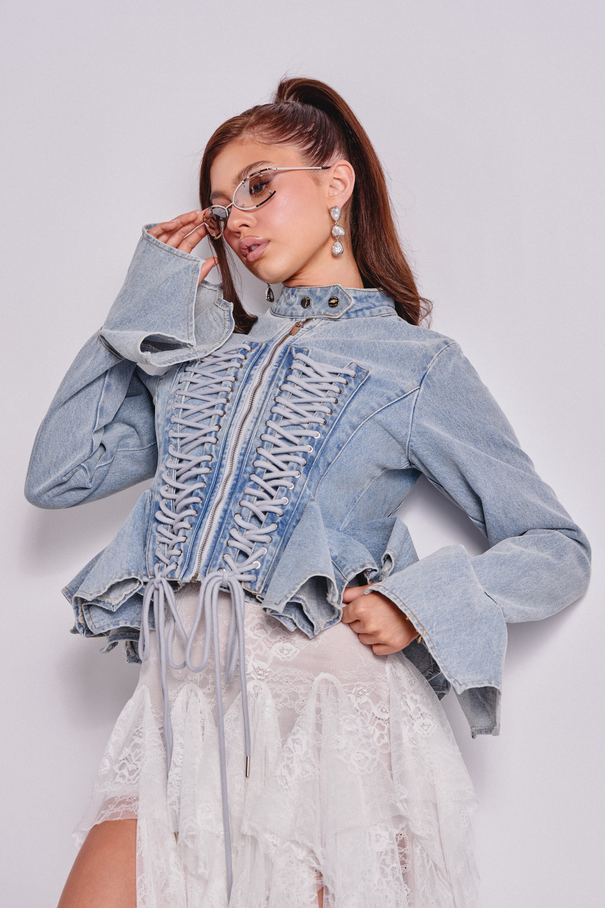 A woman poses in the DOUBLE DARE YOU PEPLUM DENIM JACKET, featuring a corseted lace-up front and flared sleeves, paired with a white lace skirt. She wears glasses, her long hair in a ponytail, against a plain light background. 0