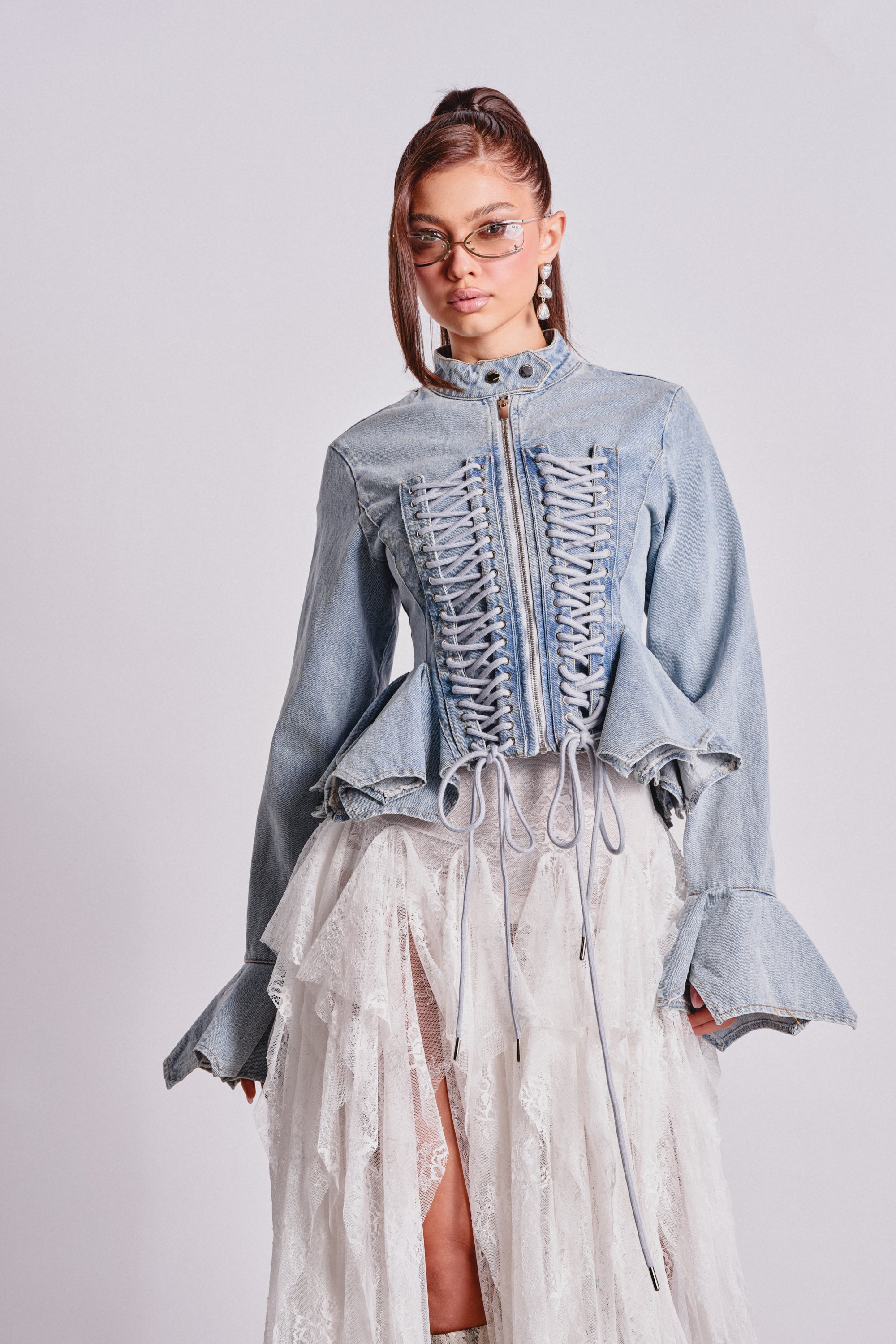 A woman in cat-eye glasses and the DOUBLE DARE YOU PEPLUM DENIM JACKET with lace-up details, paired with a layered white lace skirt, stands against a plain light background. 2