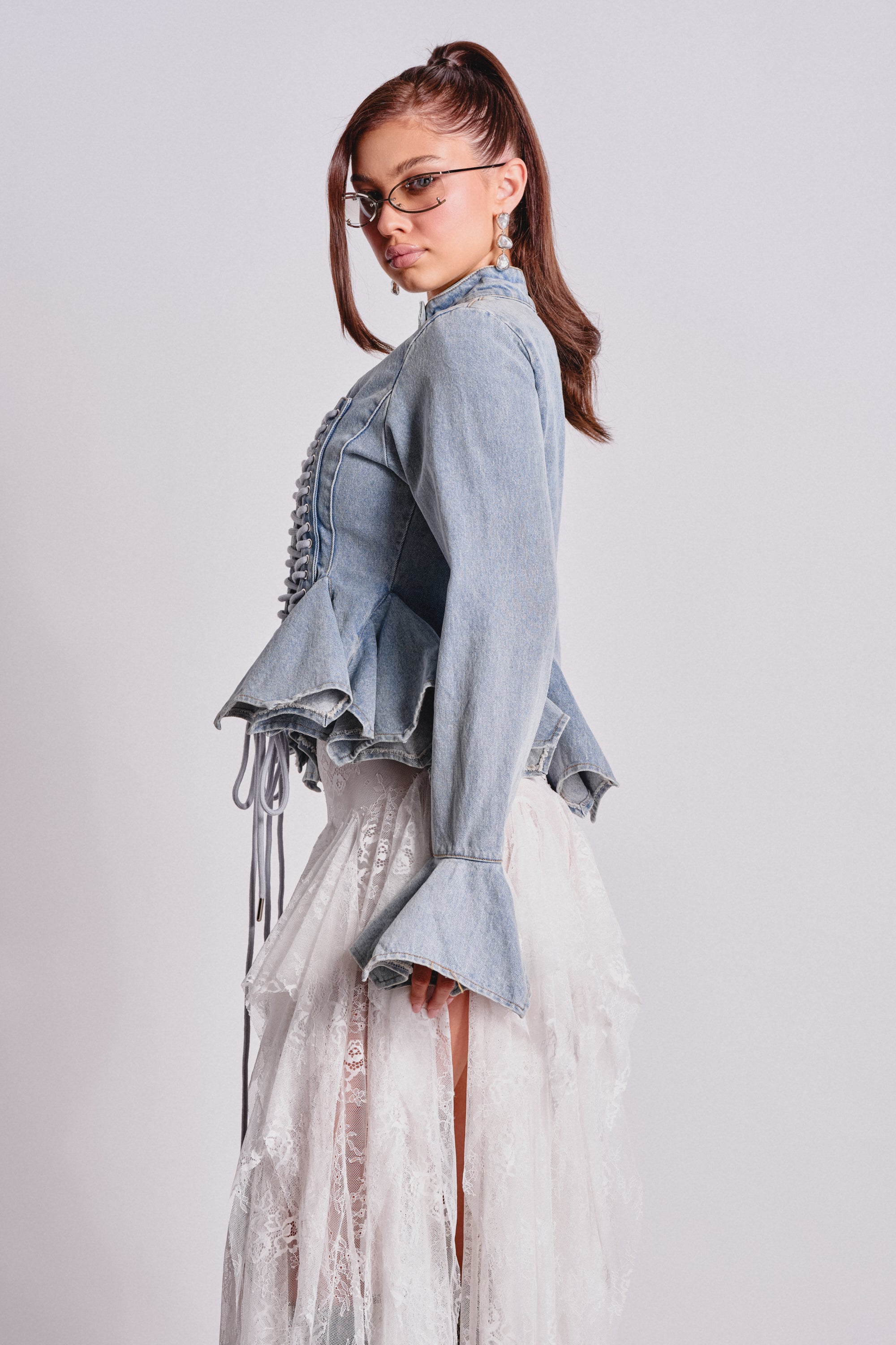 A woman in the DOUBLE DARE YOU PEPLUM DENIM JACKET, featuring a corseted fit and flared sleeves, pairs it with a white lace skirt. Her high ponytail, small sunglasses, and statement earrings complete this bold night-out look. 3