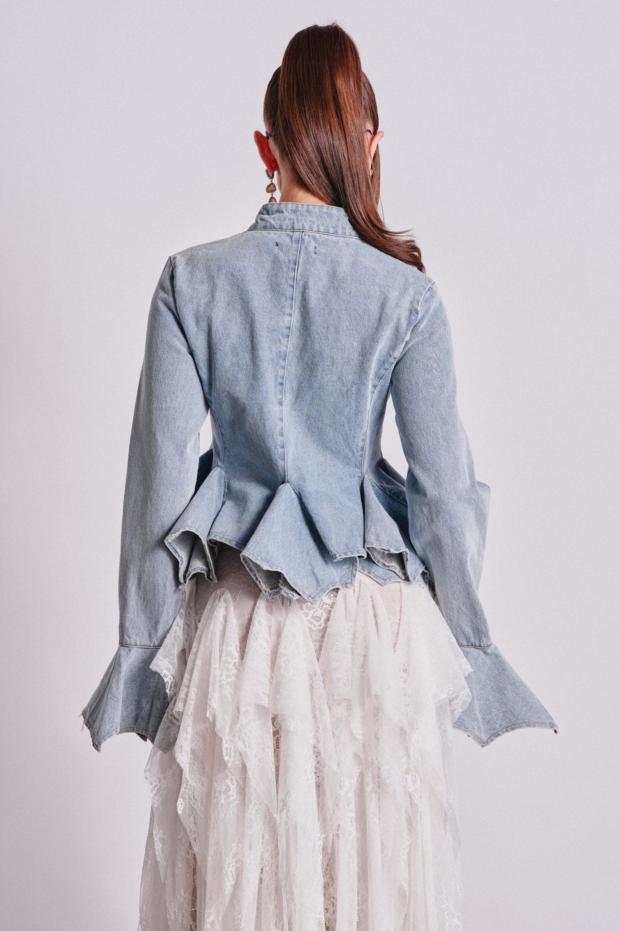 A person faces away in the DOUBLE DARE YOU PEPLUM DENIM JACKET, featuring flared cuffs, paired with a layered white lace skirt. Their long brown hair is tied back against a plain light background. 4