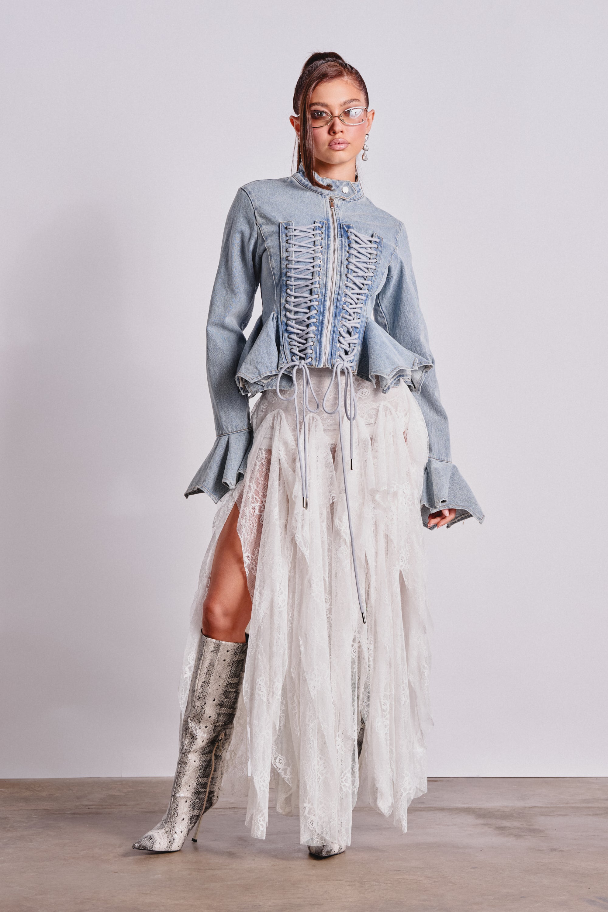 A woman poses against a plain background wearing the DOUBLE DARE YOU PEPLUM DENIM JACKET, styled with a flowing white lace skirt, silver knee-high boots, clear glasses, and her hair in a high ponytail. 5