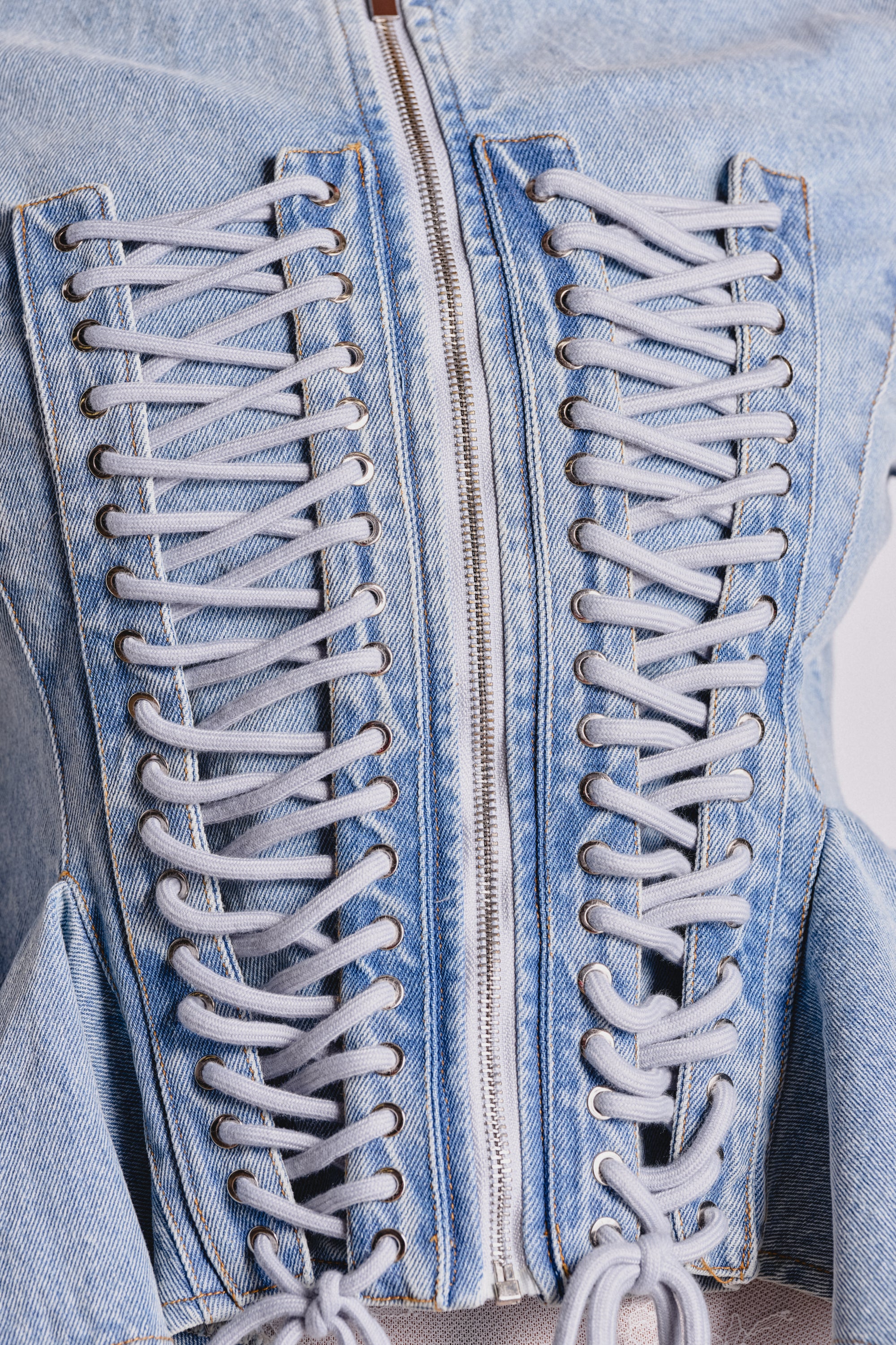 Close-up of the DOUBLE DARE YOU PEPLUM DENIM JACKET in light blue denim, featuring a central zipper and striking white lace-up panels on each side for a bold statement night out look. 6