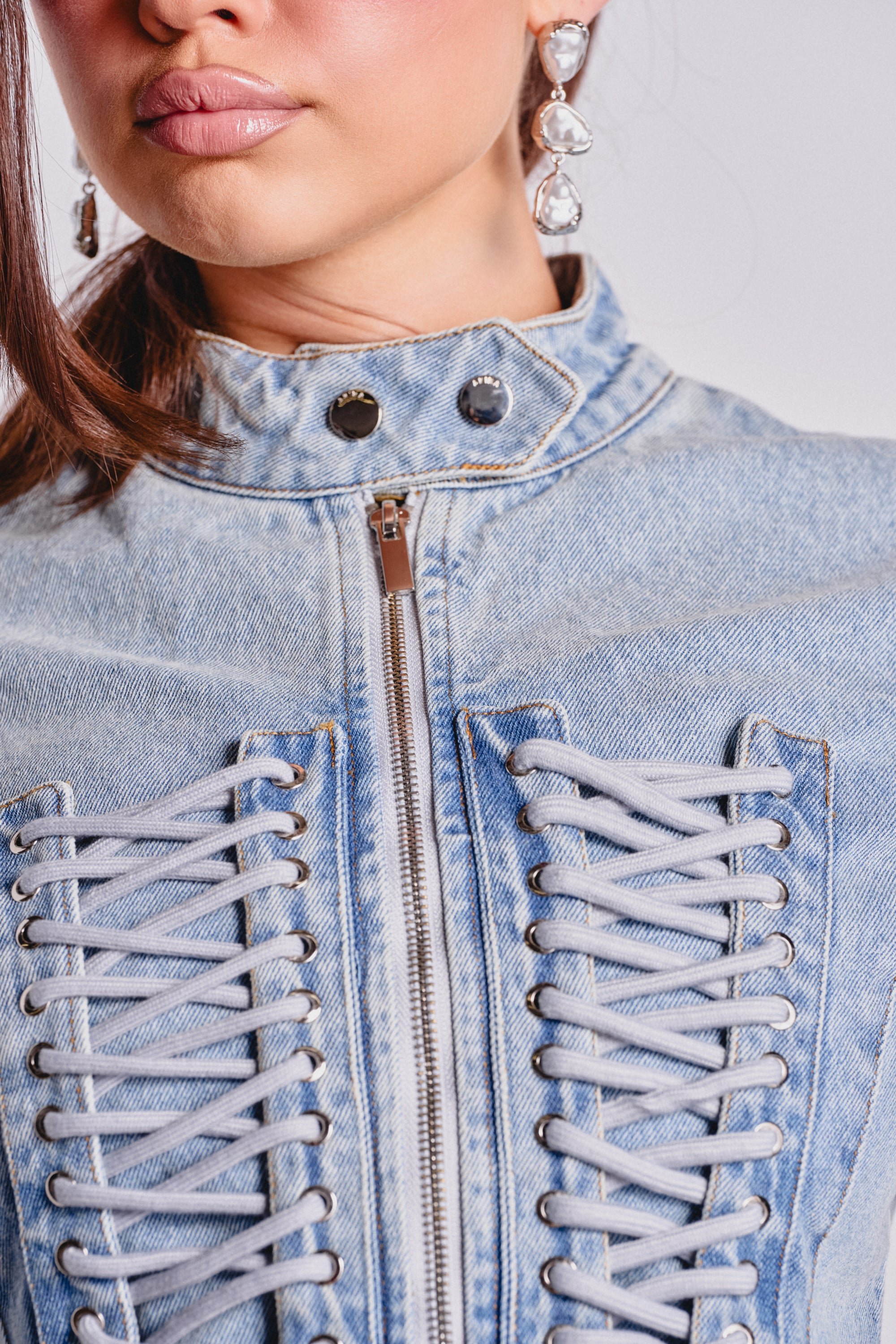 Close-up of a person wearing the DOUBLE DARE YOU PEPLUM DENIM JACKET—a light blue, corseted piece with a front zipper and crisscross lacing. Their shiny lips and heart-shaped earrings make this jacket perfect for a bold night-out look. 7