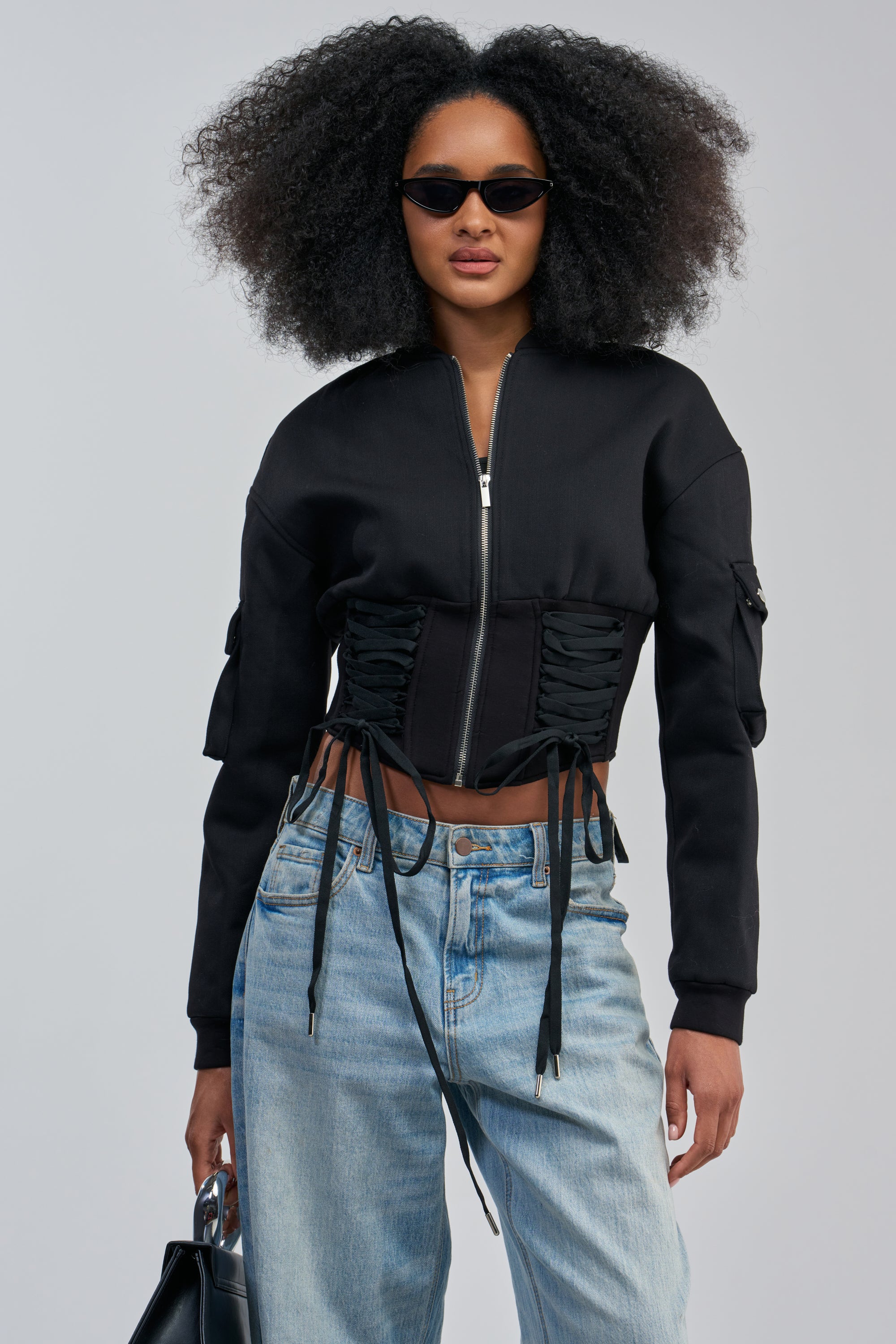 A woman with voluminous curly hair wears black sunglasses and the VIVIENNE FITTED BOMBER—a streetwear-inspired black jacket with lace-up details—paired with light blue jeans. She holds a black bag and stands against a plain light background.