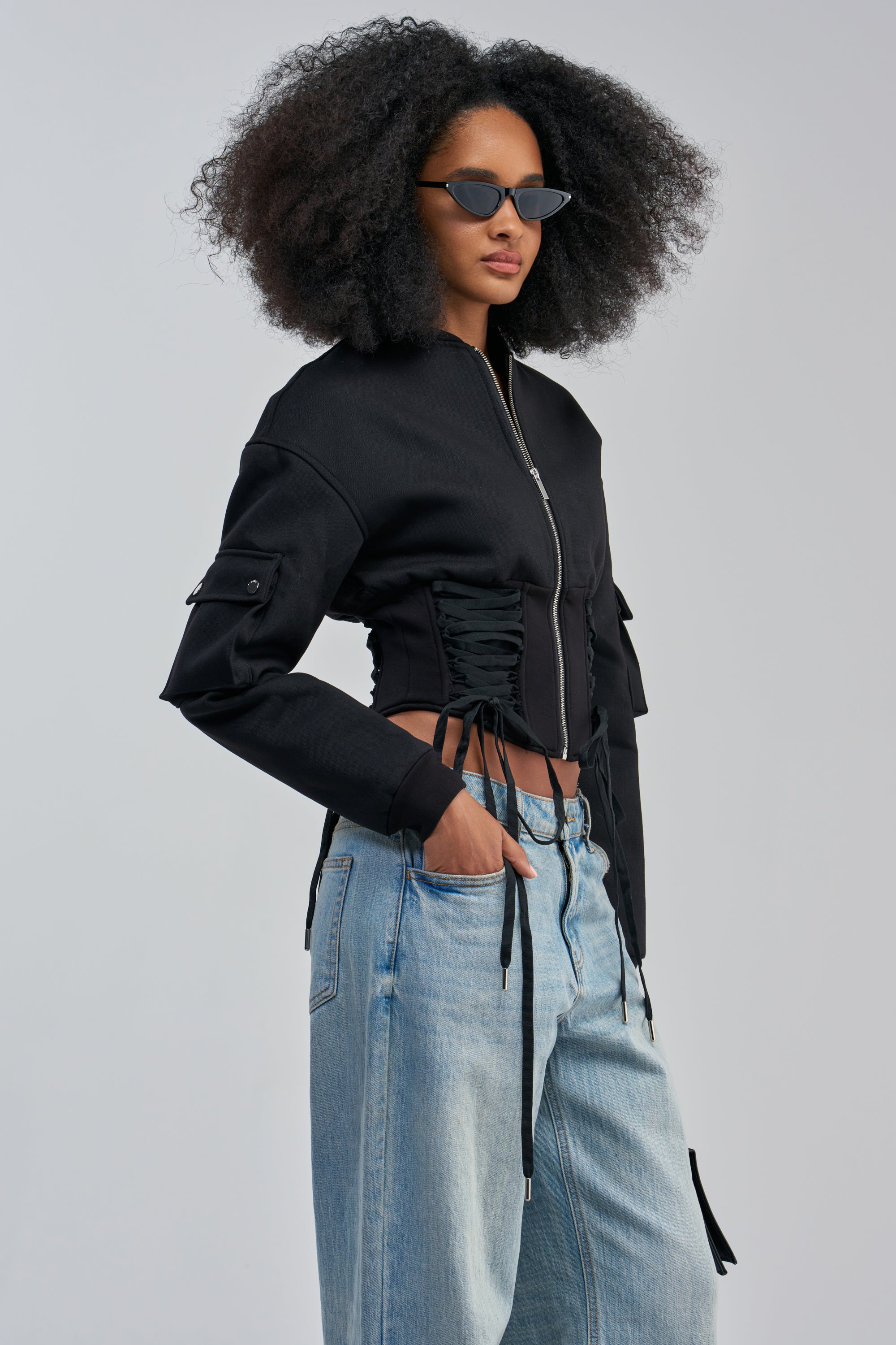 A woman with voluminous curly hair wears dark sunglasses and the VIVIENNE FITTED BOMBER styled in streetwear fashion, paired with light blue wide-leg jeans featuring cargo pockets, standing against a plain light background.