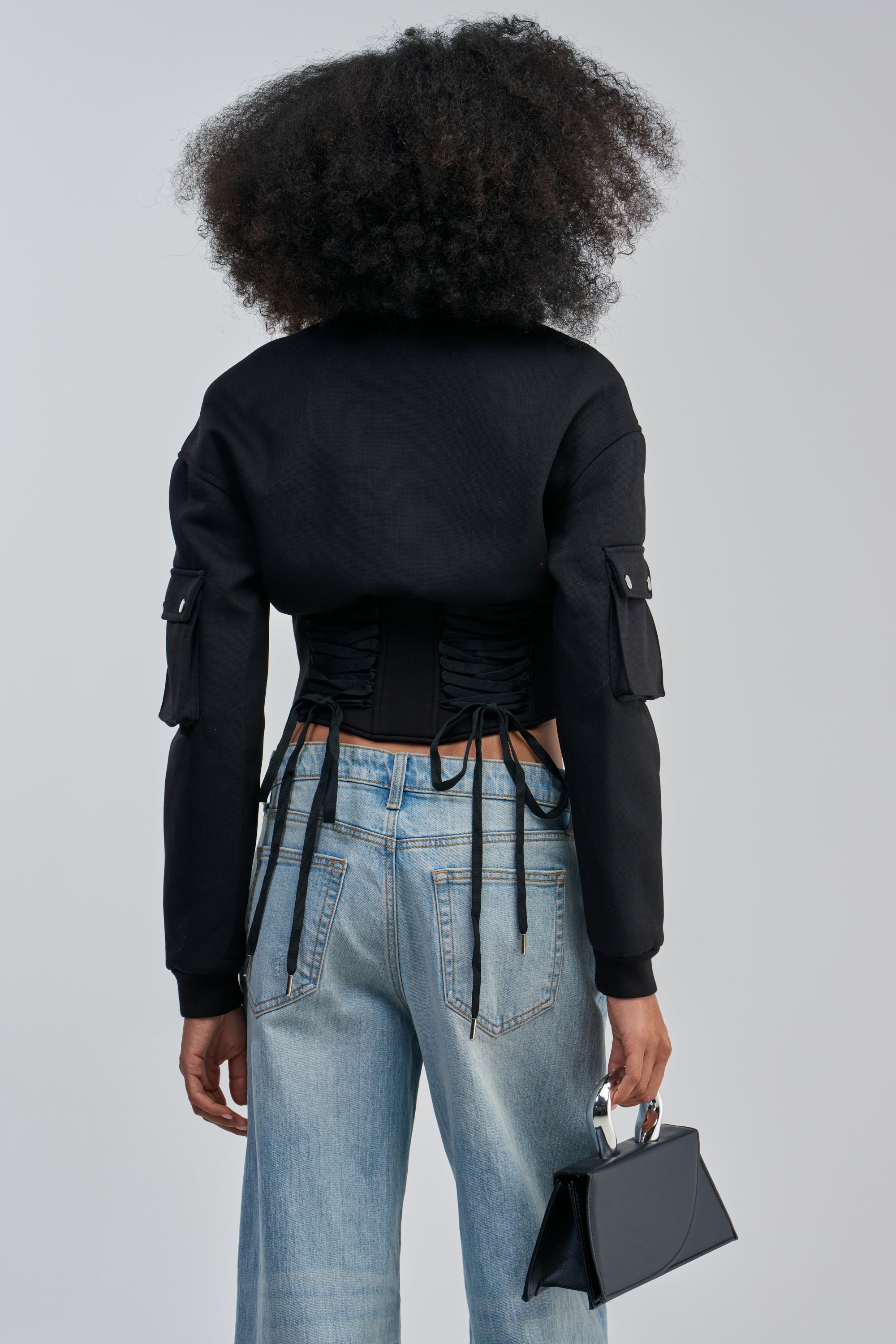 Person with a large afro hairstyle, seen from behind, wearing the VIVIENNE FITTED BOMBER with side ties and blue jeans, holding a small black handbag in their left hand—a bold streetwear look.