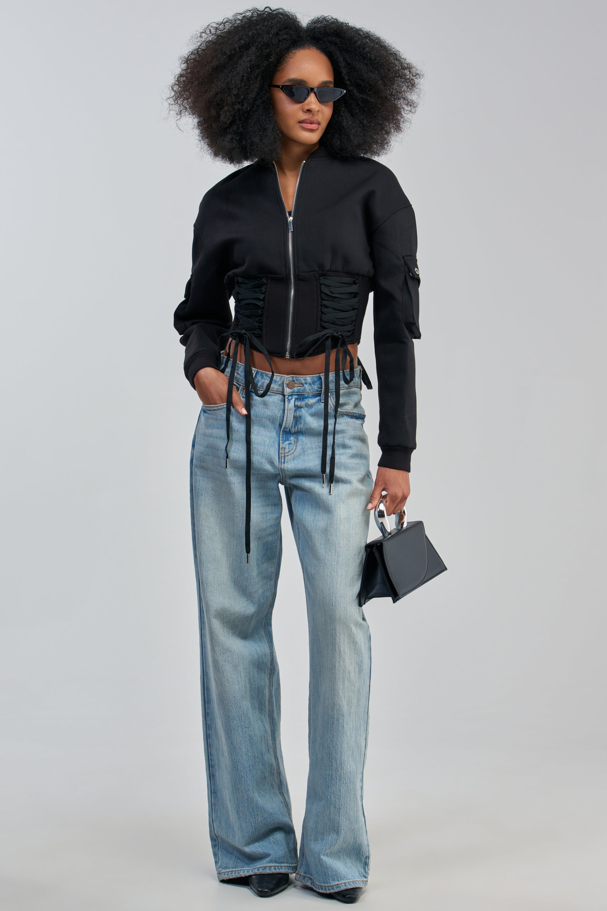 A woman with curly hair wears cat-eye sunglasses, a VIVIENNE FITTED BOMBER styled in streetwear fashion, light blue wide-leg jeans, and holds a small black handbag.