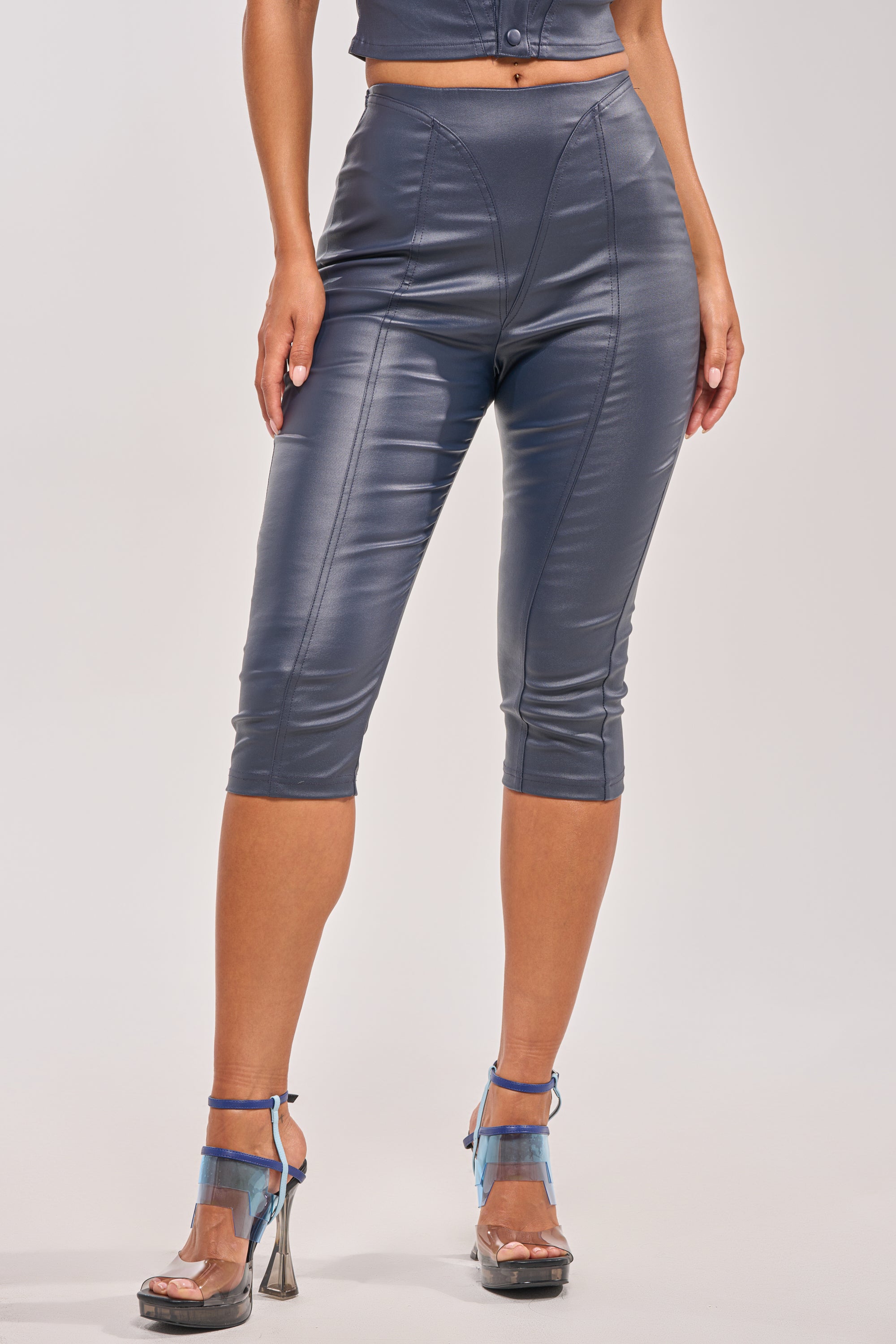 A woman wears the BIG CITY LIVIN FAUX LEATHER CAPRI in shiny gray, paired with clear high-heeled sandals with blue ankle straps. She stands against a plain light background, with only her lower half visible. 1