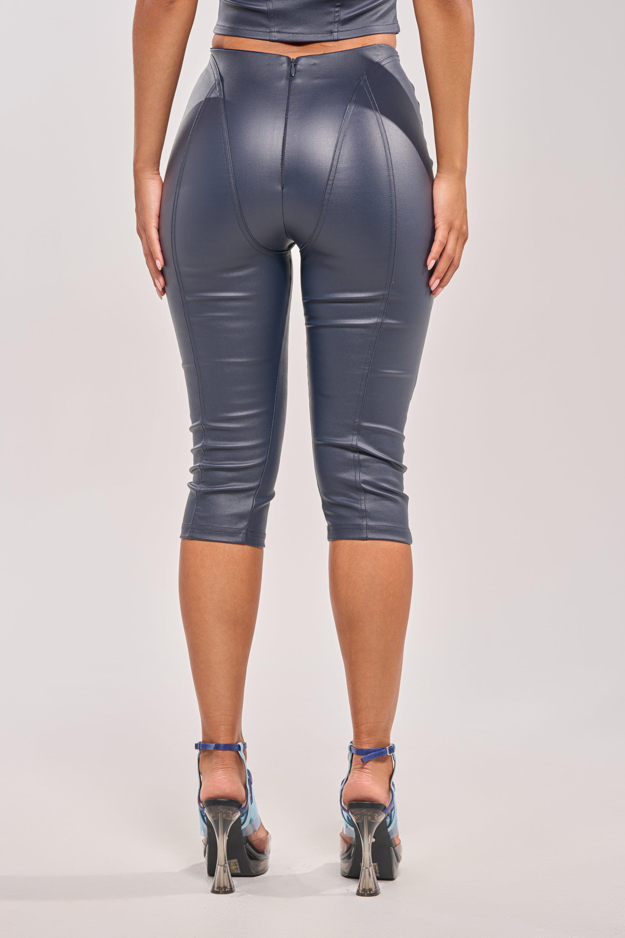 A person is seen from behind wearing the BIG CITY LIVIN FAUX LEATHER CAPRI—navy, high-waisted, curve-hugging faux leather pants with visible seam detail and a shiny finish—paired with strappy high heels and ending just below the knees. 3