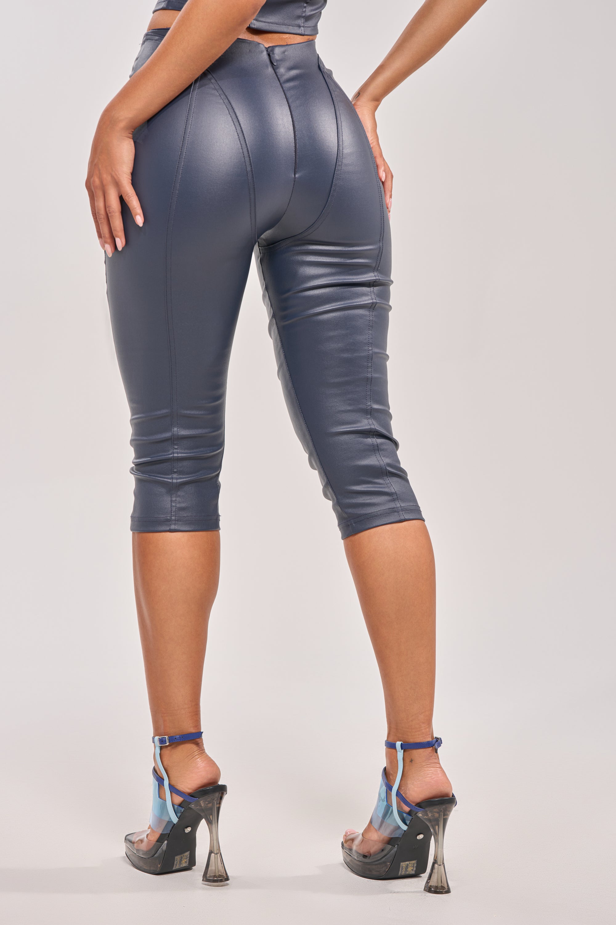 Wearing the BIG CITY LIVIN FAUX LEATHER CAPRI in shiny navy with a curve-hugging fit and transparent high-heeled sandals, a model poses against a plain background, shown from the back with one hand on her hip. 4