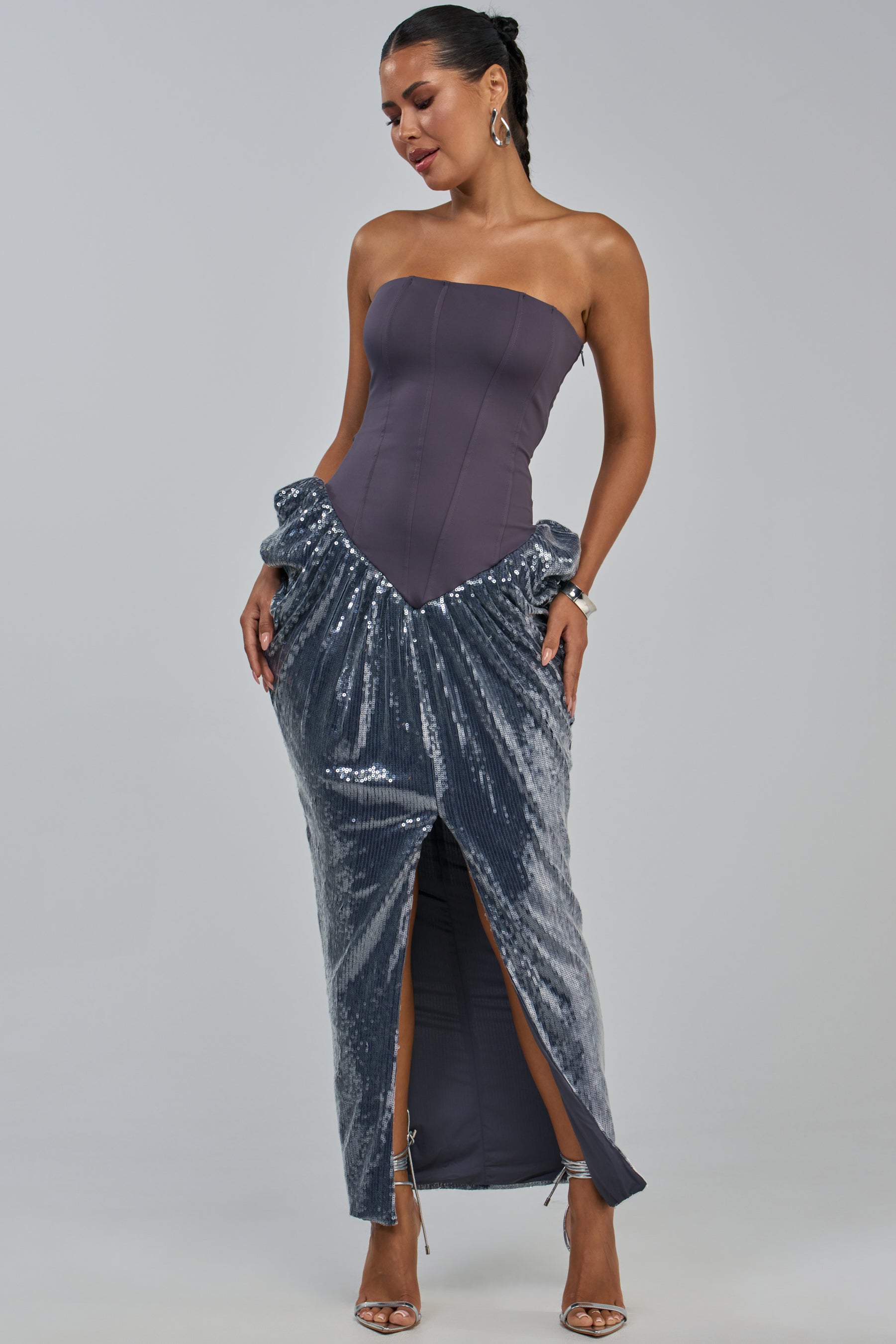 Wearing the GLOW GETTER SEQUIN CORSET MAXI DRESS IN CHARCOAL, a woman stands in heels, looking down with a slight smile. The strapless, fitted corset dress features a shiny, textured sequin skirt and a front slit.