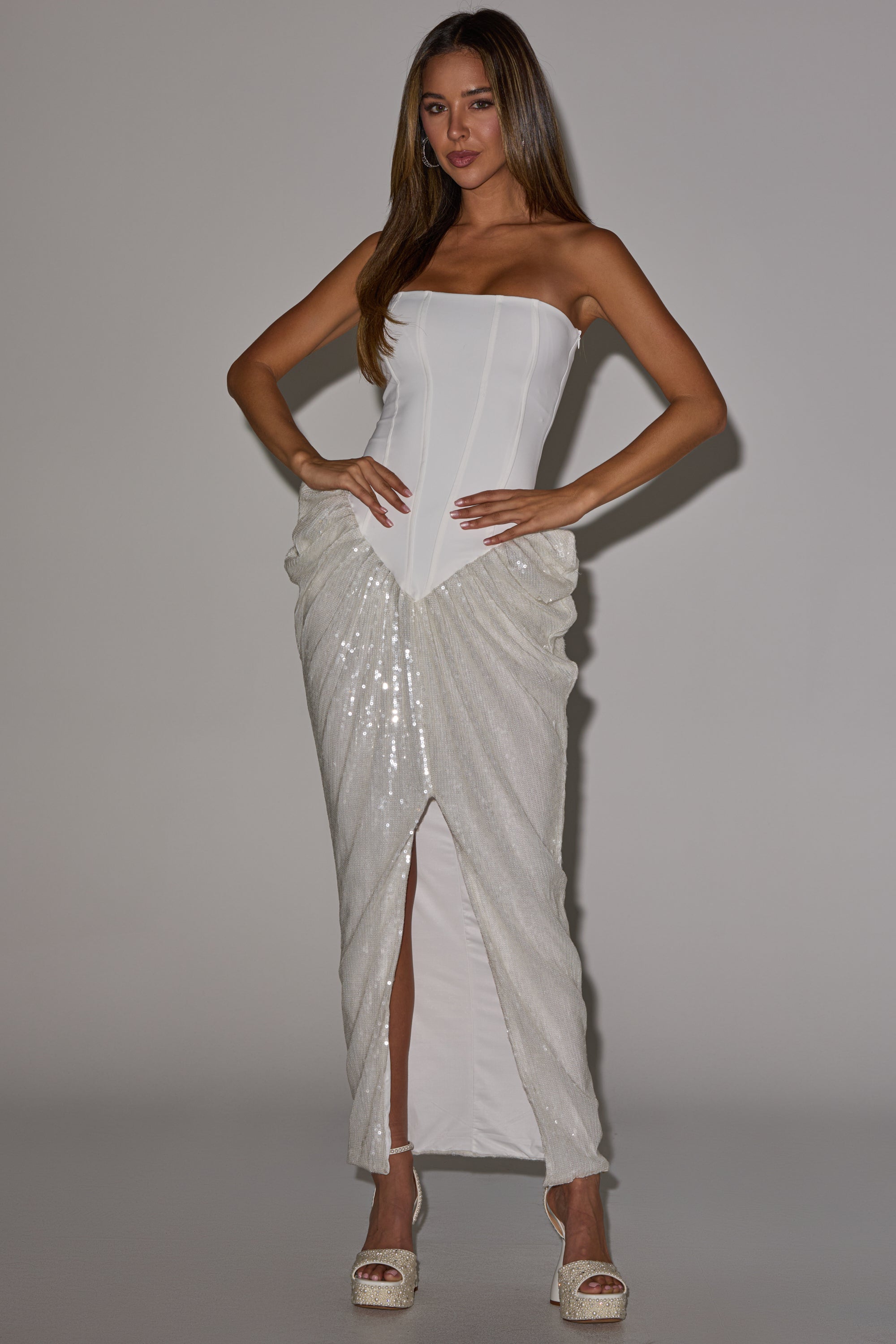 A woman models the GLOW GETTER SEQUIN CORSET MAXI DRESS—a strapless white maxi with a fitted bodice, shimmering ruched skirt, and front slit—paired with white platform heels. She stands against a plain, light backdrop with straight hair.