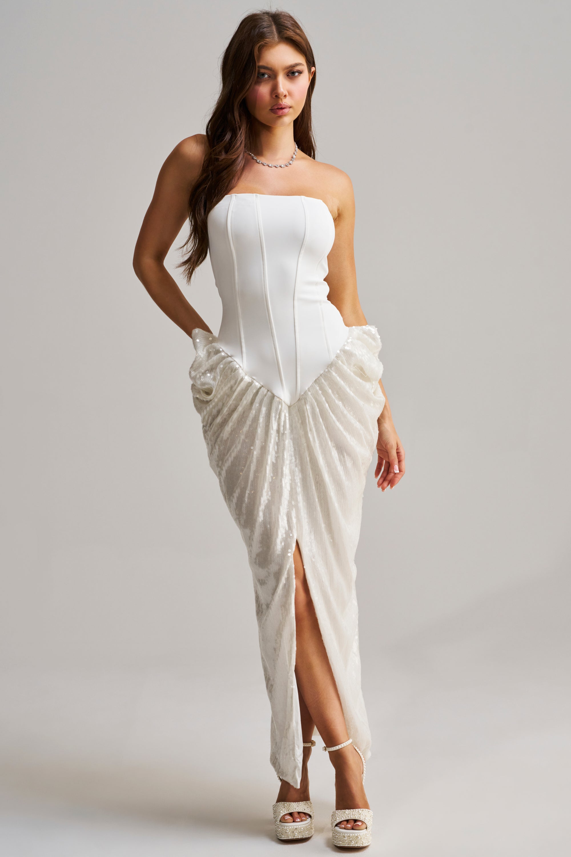 Wearing the GLOW GETTER SEQUIN CORSET MAXI DRESS with a structured, strapless bodice and a draped skirt with a front slit, she pairs platform heels and a necklace. Her long wavy brown hair completes the chic look against a plain background.