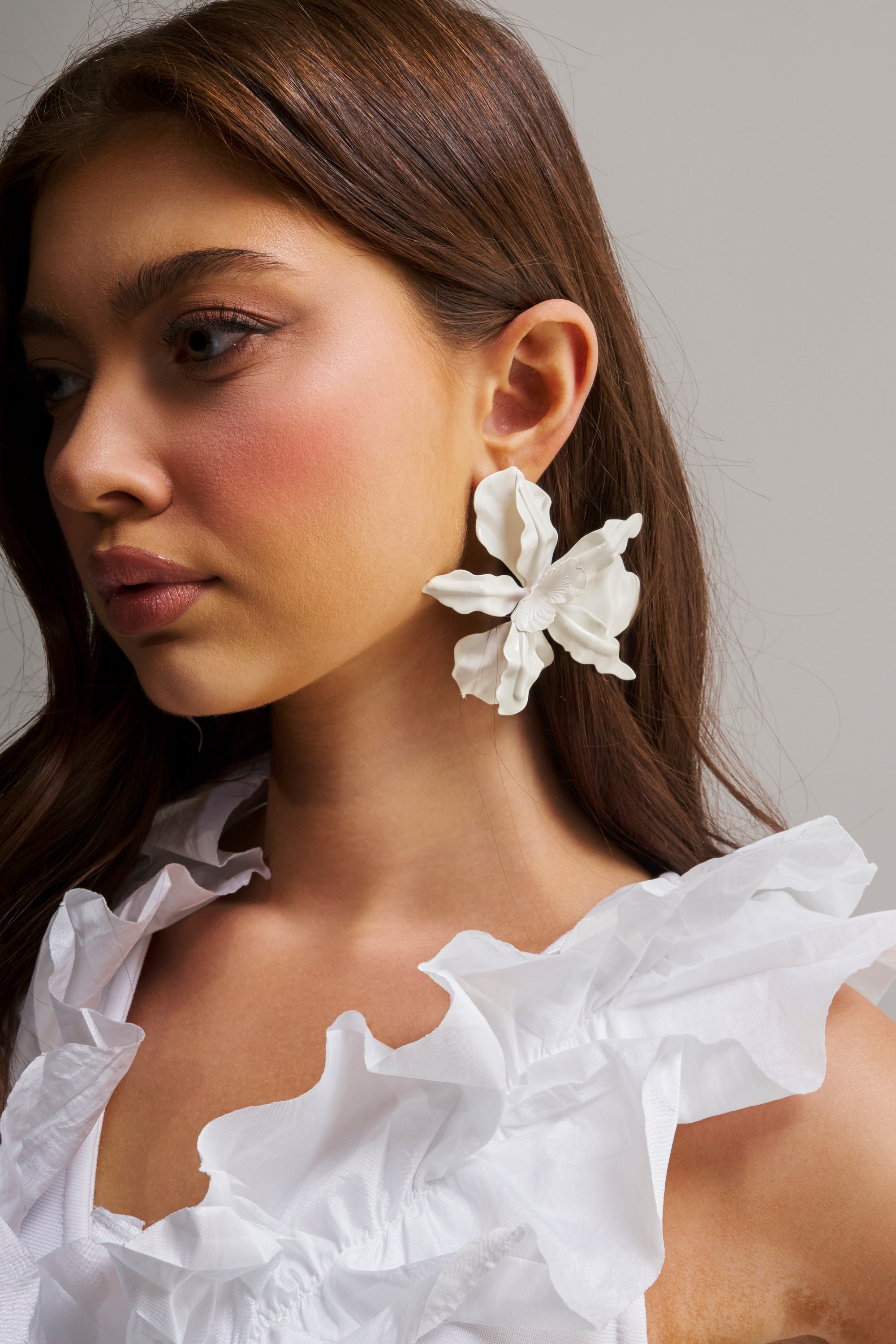 A woman with long brown hair wears a white ruffled top and the NEVER SETTLE EARRING, a large abstract floral statement piece, against a plain neutral background.