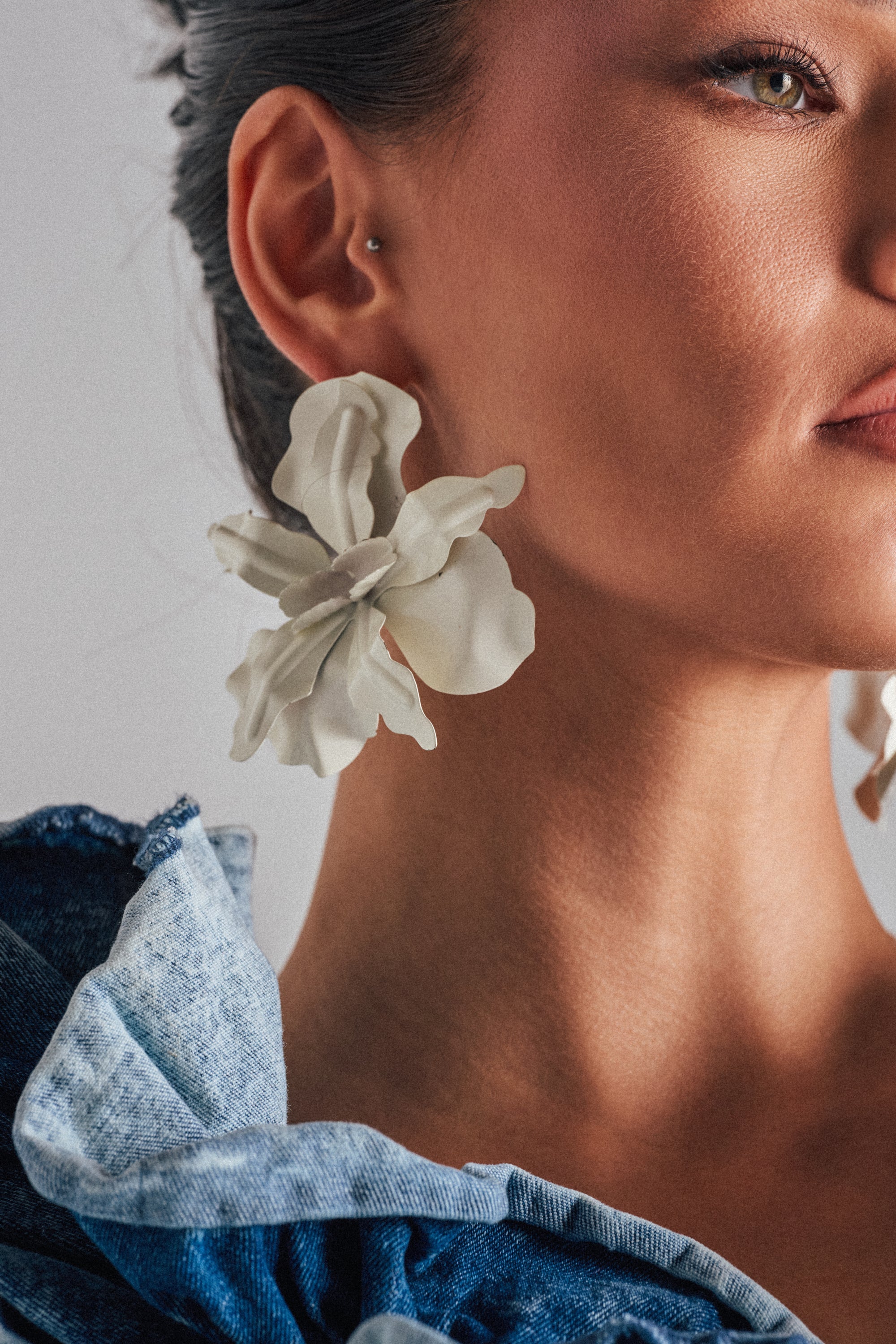 A close-up of a woman wearing large white floral NEVER SETTLE EARRINGs and a blue ruffled top, showing only the lower half of her face, ear, neck, and part of her shoulder. 0