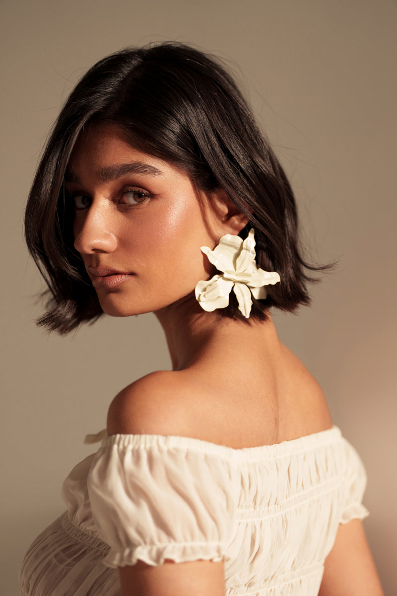 A woman with short dark hair and glowing skin wears a white off-the-shoulder top and the NEVER SETTLE EARRING, gazing softly over her shoulder against a neutral background. 2