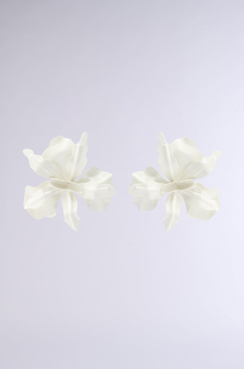 Two white, delicate flower-shaped NEVER SETTLE EARRINGS with ruffled petals are centered on a plain, light gray background. Their symmetrical design and sculpted floral form evoke the elegance of modern jewelry. 4
