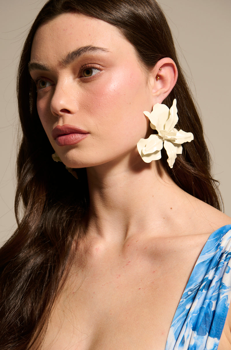 A woman with long brown hair wears the NEVER SETTLE EARRING, featuring a floral post. She pairs them with a blue patterned top and natural makeup, gazing to the side against a neutral background. 5