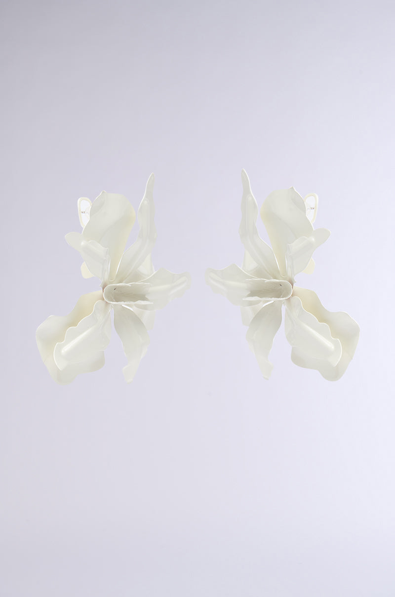 The NEVER SETTLE EARRING features two white, flower-shaped earrings with layered petals, a metallic base, and a smooth matte finish. Their modern sculptural floral design is displayed against a plain light gray background. 6