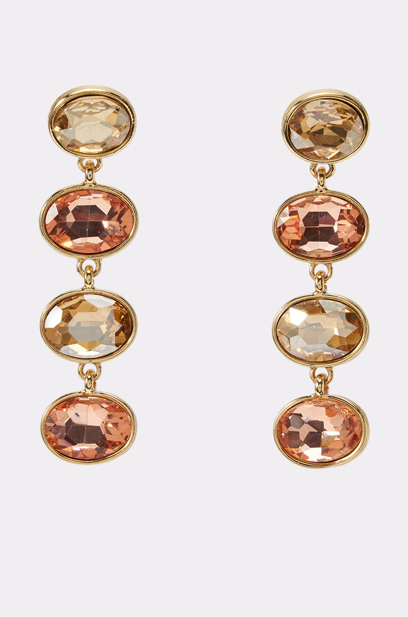 Introducing the GOLDEN HOUR DRIP EARRINGS: Gold-tone drop earrings featuring three vertically-set, oval-shaped faceted gemstones in shimmering gold, champagne, and rose hues. Set in delicate bezel settings against a light backdrop, these elegant rhinestone earrings infuse any outfit with a refined touch of radiant sparkle.