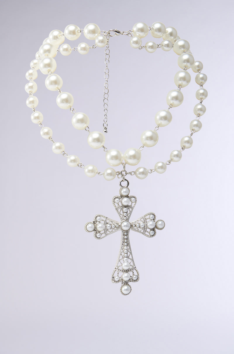 The LIKE A PRAYER PEARL NECKLACE features an elegant dual-layer design with two rows of pearls linked by a delicate chain. It boasts a silver cross pendant adorned with tiny pearls and intricate details, set against a plain gray background for timeless allure.