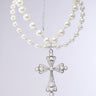 The LIKE A PRAYER PEARL NECKLACE features an elegant dual-layer design with two rows of pearls linked by a delicate chain. It boasts a silver cross pendant adorned with tiny pearls and intricate details, set against a plain gray background for timeless allure.